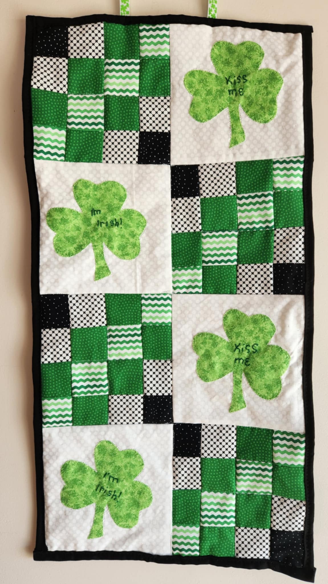 A quilted wall hanging with a St. Patrick's Day theme featuring green shamrocks and text that reads 'Kiss Me, I'm Irish'. With a patchwork patters of black, white and greens