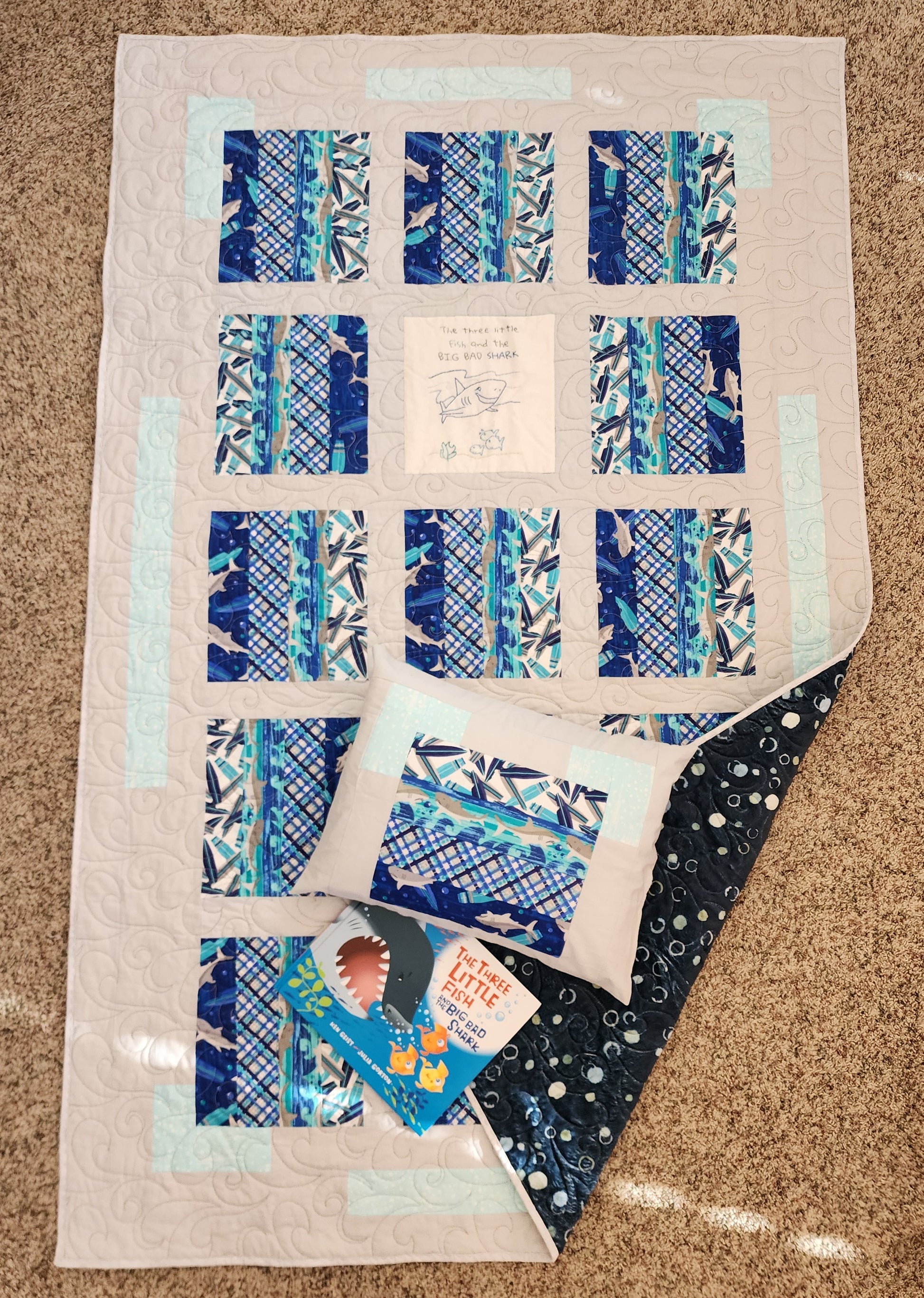 A shark-themed quilt with blue and gray patterns laid out on a flat surface with a pillow with the same pattern and a children's book. Bottom corner is turned up showing blue with dots out of mink