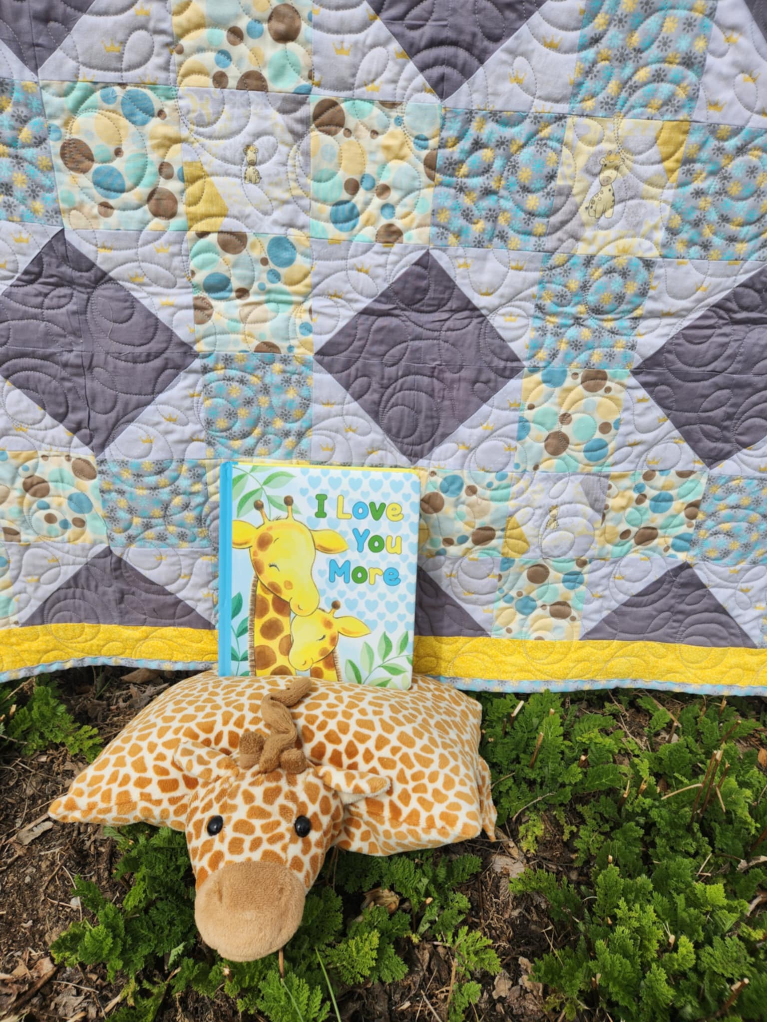 A Quilt With a octagon patters with squares of circles in brown, blue and yellow, rectangles of solid dark gray and light gray background with gold crowns and a square with a giraffe in the center surrounded by gray and gold octagons. with a yellow trim. A stuff giraffe and a book called I love you more at the bottom