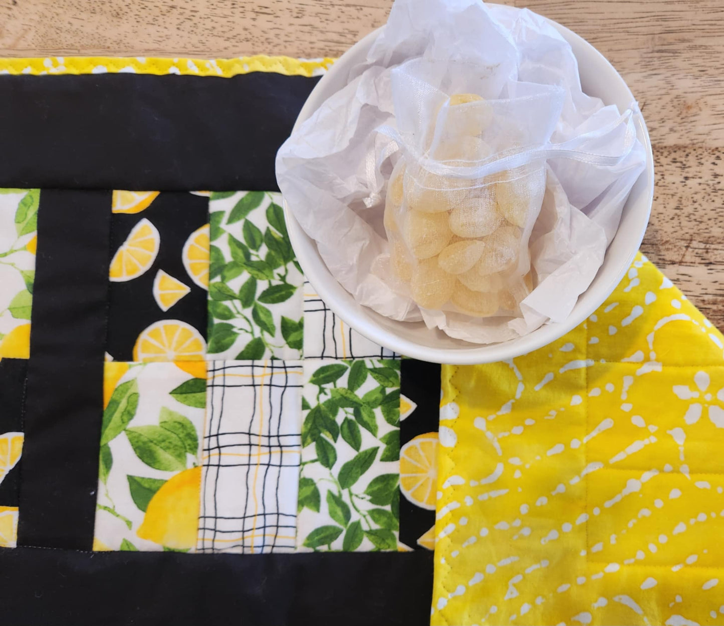 close up of table runner with Yellow and white binding, with black surrounding four square with the pattern of Lemons with green leaves fabric, black background with sliced lemons fabric, green leaves with white background fabric, squiggly black and yellow lines with white back round fabric, repeated twice for each square. The right corner is turned down showing yellow fabric with white flowers. There is a bowl with lemons and greens on it.