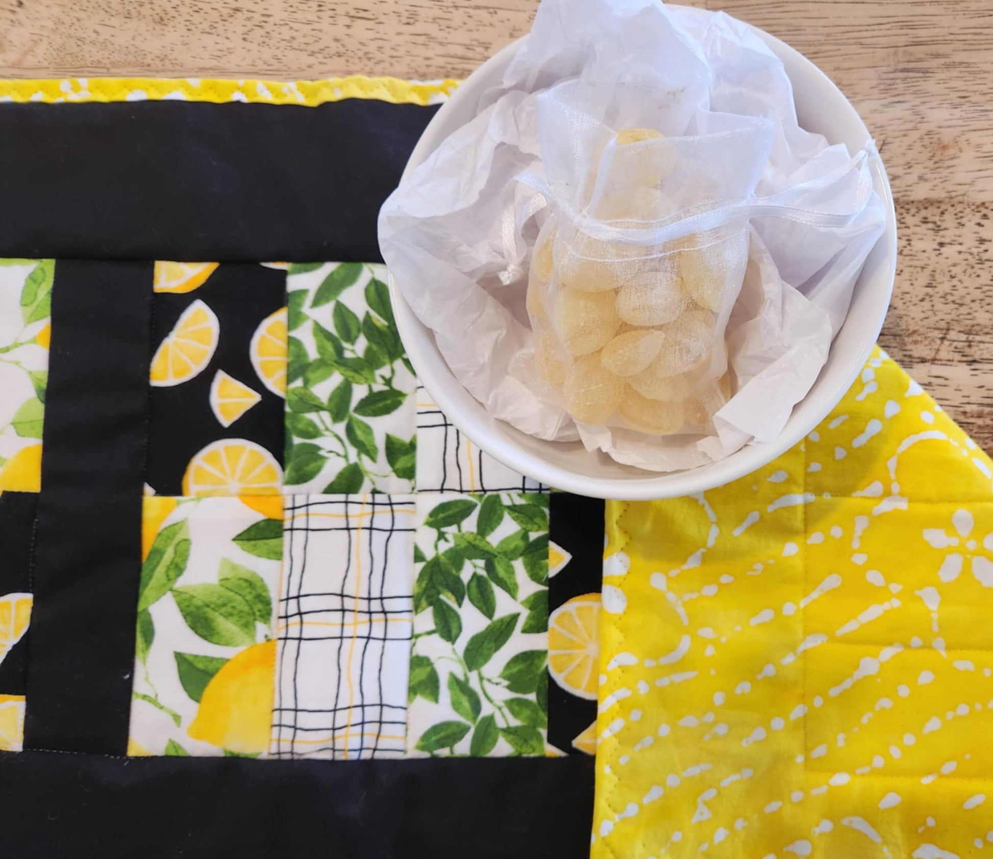 close up of table runner with Yellow and white binding, with black surrounding four square with the pattern of Lemons with green leaves fabric, black background with sliced lemons fabric, green leaves with white background fabric, squiggly black and yellow lines with white back round fabric, repeated twice for each square. The right corner is turned down showing yellow fabric with white flowers. There is a bowl with lemons and greens on it.