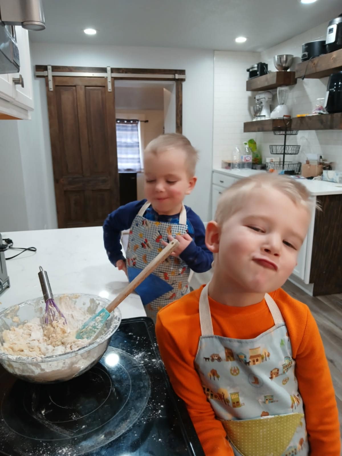 Two children wearing western aprons, one has a small western town with a blue background. Has a pocket that is a tan fabric. The second one has cowboy boots and hats with a blue and white checkered as the background. They are standing in a kitchen environment, with one child stirring ingredients in a bowl