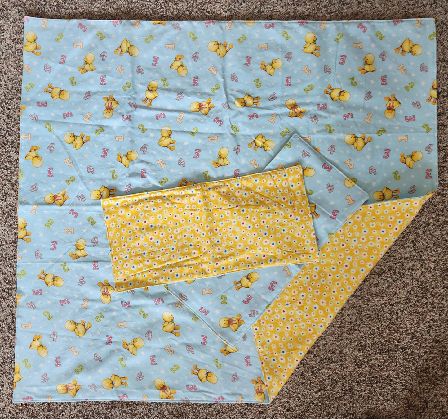 Square Receiving Blanket with Baby ducks and numbers pattern with blue background Fleece. With corner turned down showing colorful dots with yellow background fleece. With two burp rags made from the same fabric on top of the blanket