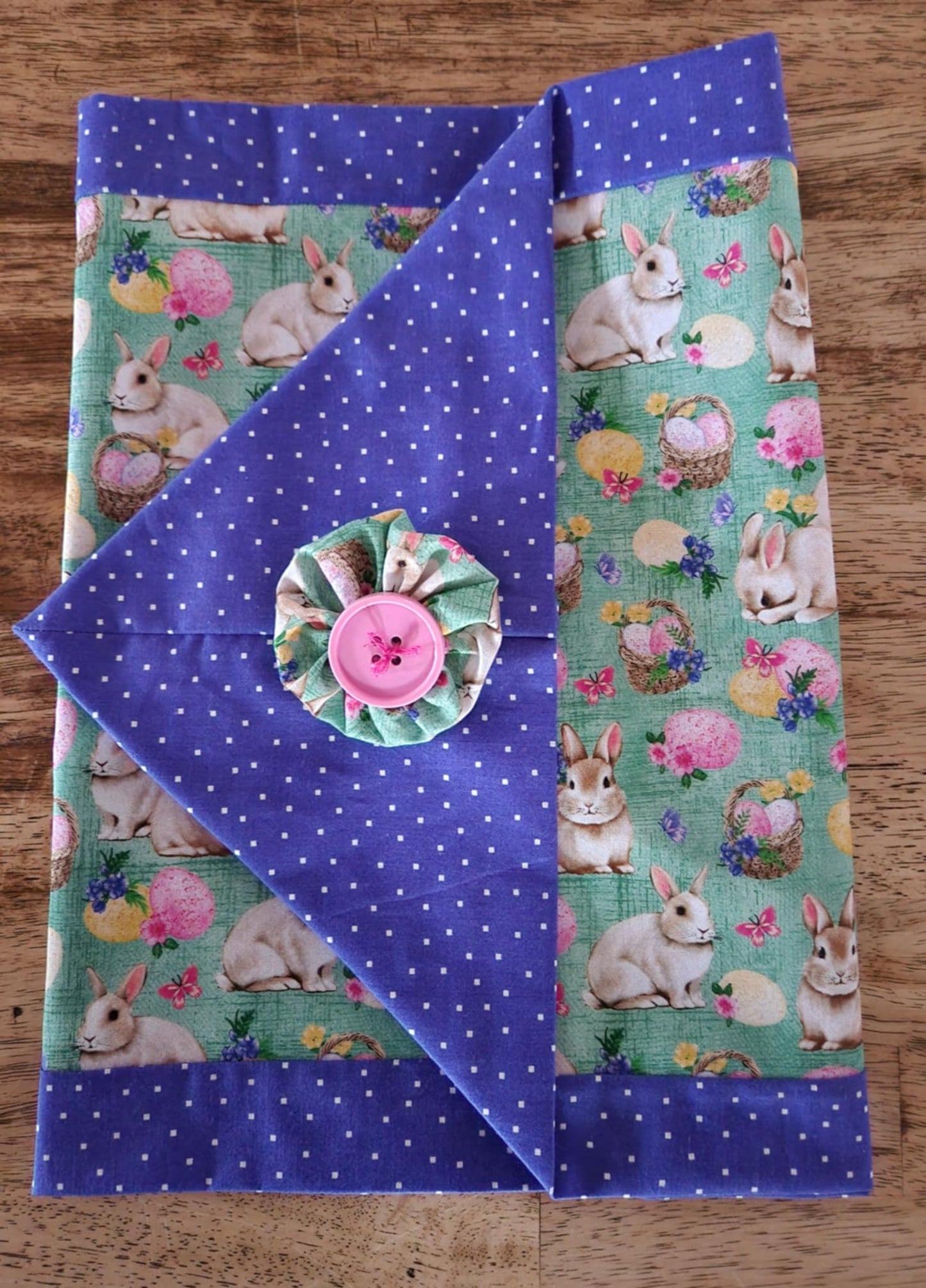 A Folded table runner with bunnies' with baskets filled with Easter eggs  in the middle surrounded with a purple poke-a-dots outside with a pink button