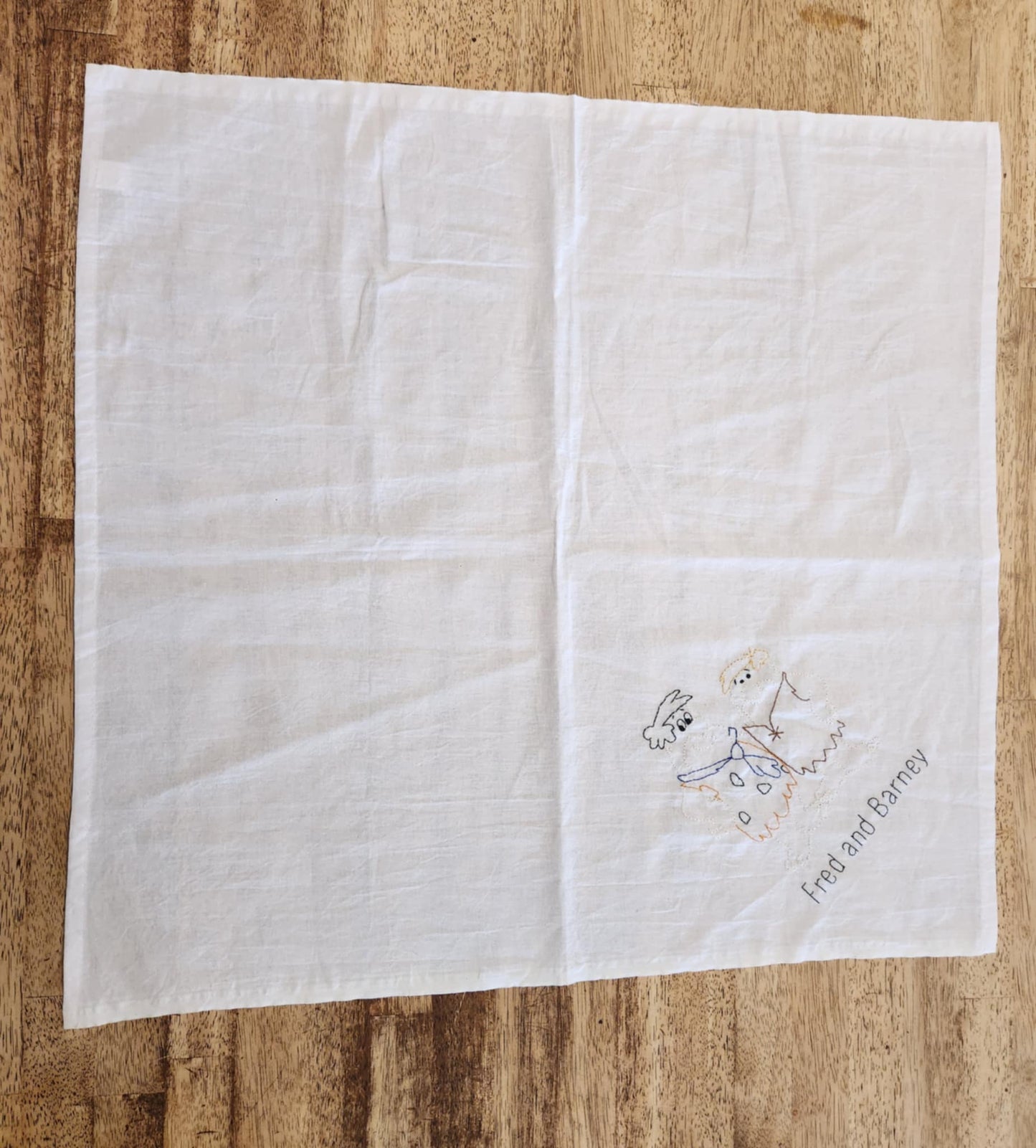 A white square towel made of cotton placed on a wood surface. In the right corner is the words Fred and Barney in black thread and above that is a outlined picture of the characters in colored thread.