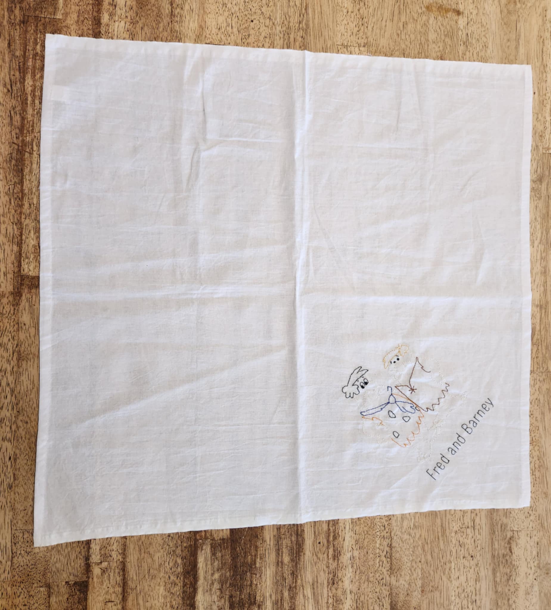 A white square towel made of cotton placed on a wood surface. In the right corner is the words Fred and Barney in black thread and above that is a outlined picture of the characters in colored thread.