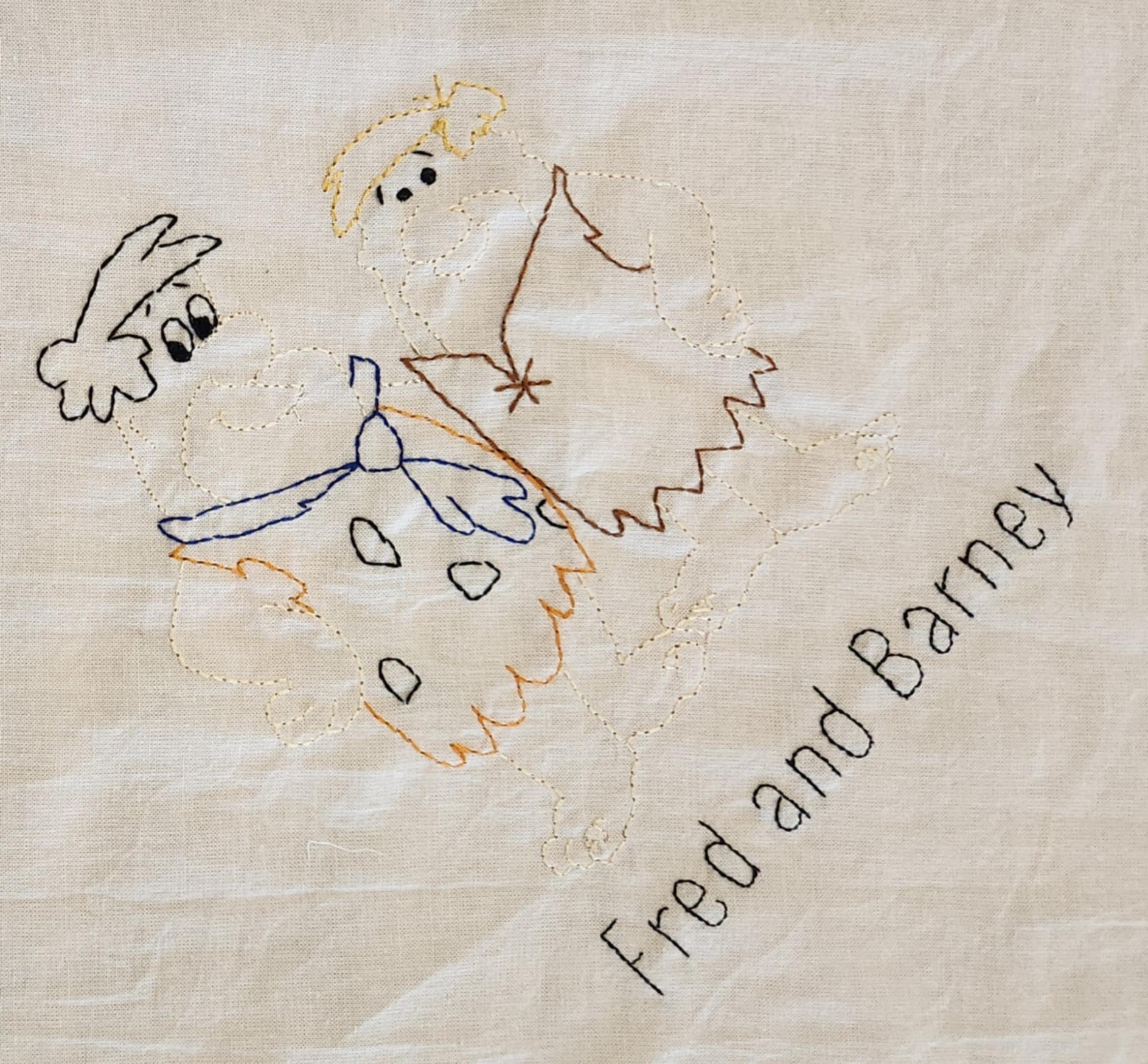 A close up on a white cotton towel With the words Fred and Barney in black thread. Above that is the image of the characters with colored thread.