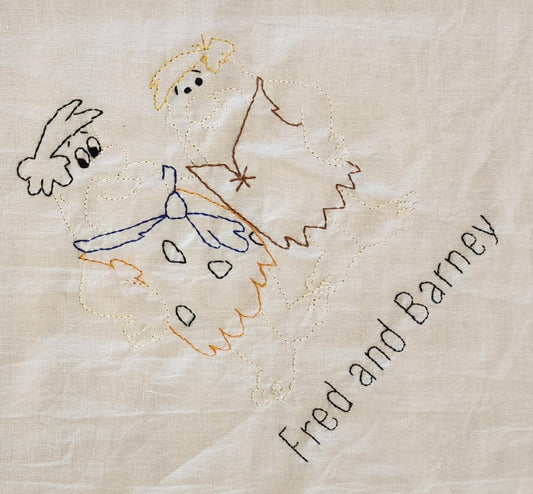 A close up on a white cotton towel With the words Fred and Barney in black thread. Above that is the image of the characters with colored thread.
