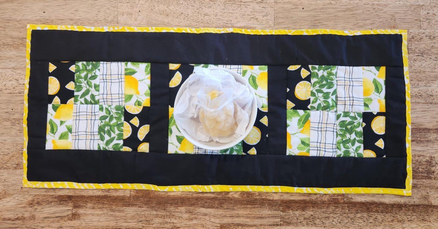 A table runner with Yellow and white binding, with black surrounding four square with the pattern of Lemons with green leaves fabric, black background with sliced lemons fabric, green leaves with white background fabric, squiggly black and yellow lines with white back round fabric, repeated twice for each square. There is a bowl with lemons and greens on it.
