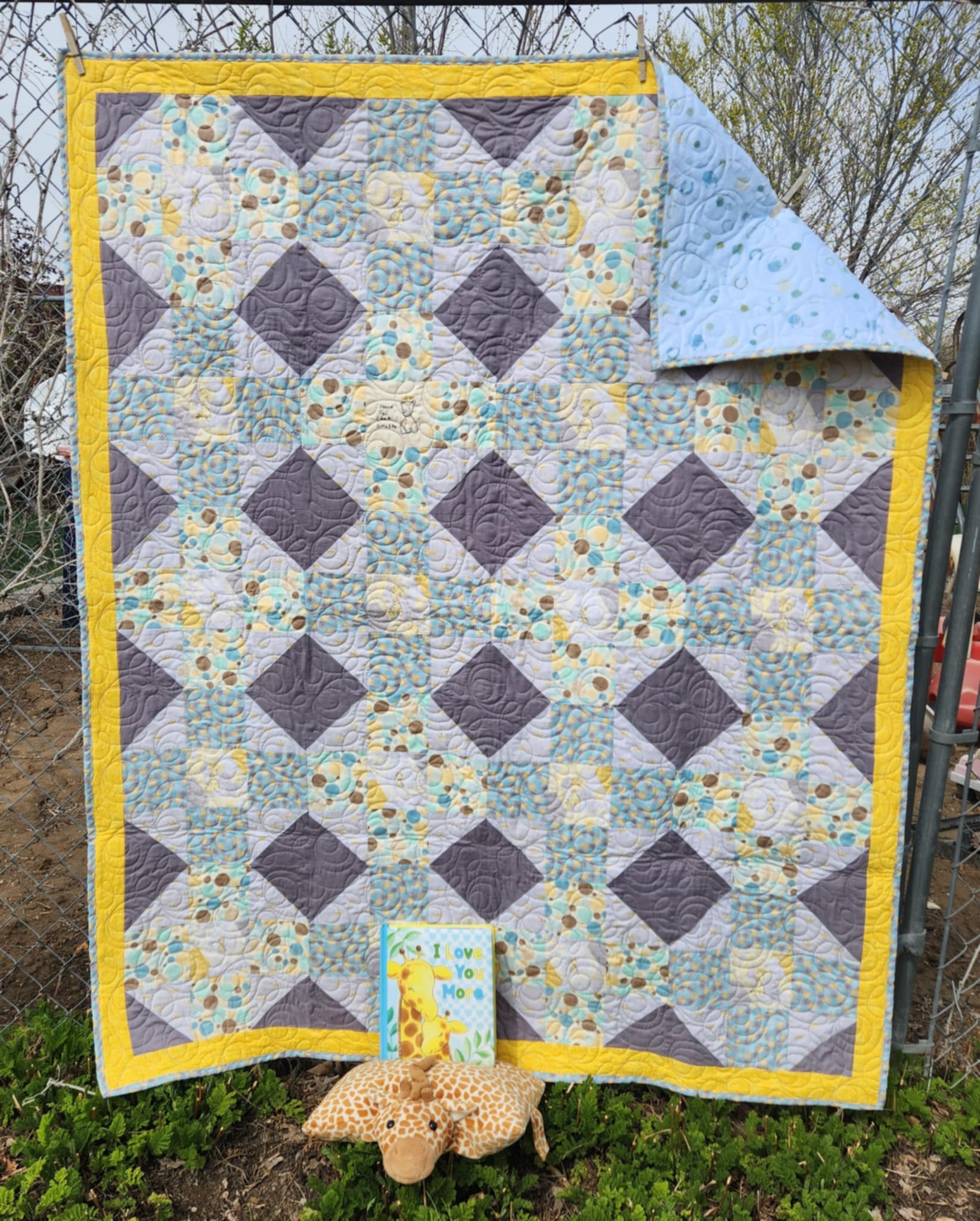 A Quilt With a octagon patters with squares of circles in brown, blue and yellow, rectangles of solid dark gray and light gray background with gold crowns and a square with a giraffe in the center surrounded by gray and gold octagons. The top right corner is turned down showing the Light blue background with full circles and half circles on minky fabric.  With a stuff giraffe and a book at the bottom