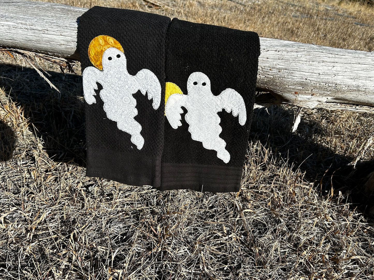 Two black hand towels, each has a white ghost with a different pattern. A moon over there left shoulder which is yellow but diffrent patterns