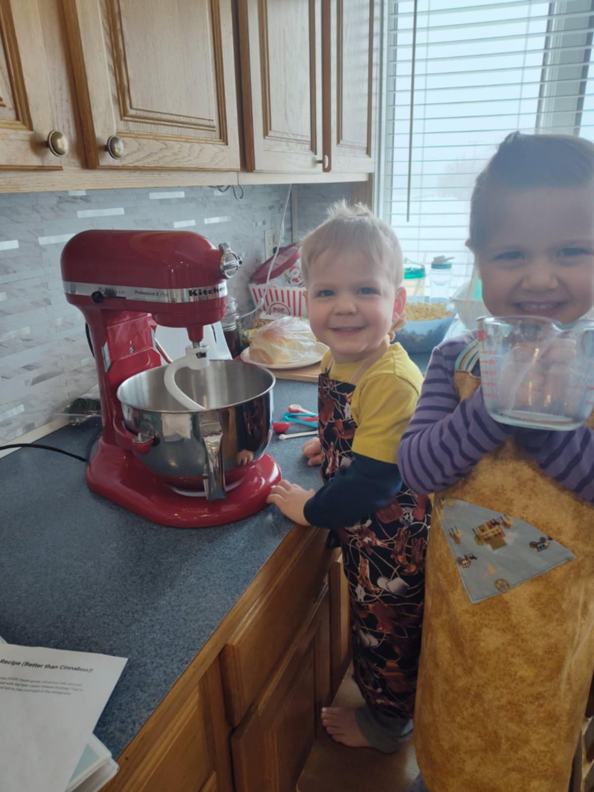 A Smiling girl wearing a light tan with dark tan spots and a sky blue pocket with a western town pattern apron holding a glass liquid measuring cup. A boy wearing a black with a western pattern of saddles boots and ropes apron smiling at the camera.  They are standing next to a counter that holds a stand mixer.