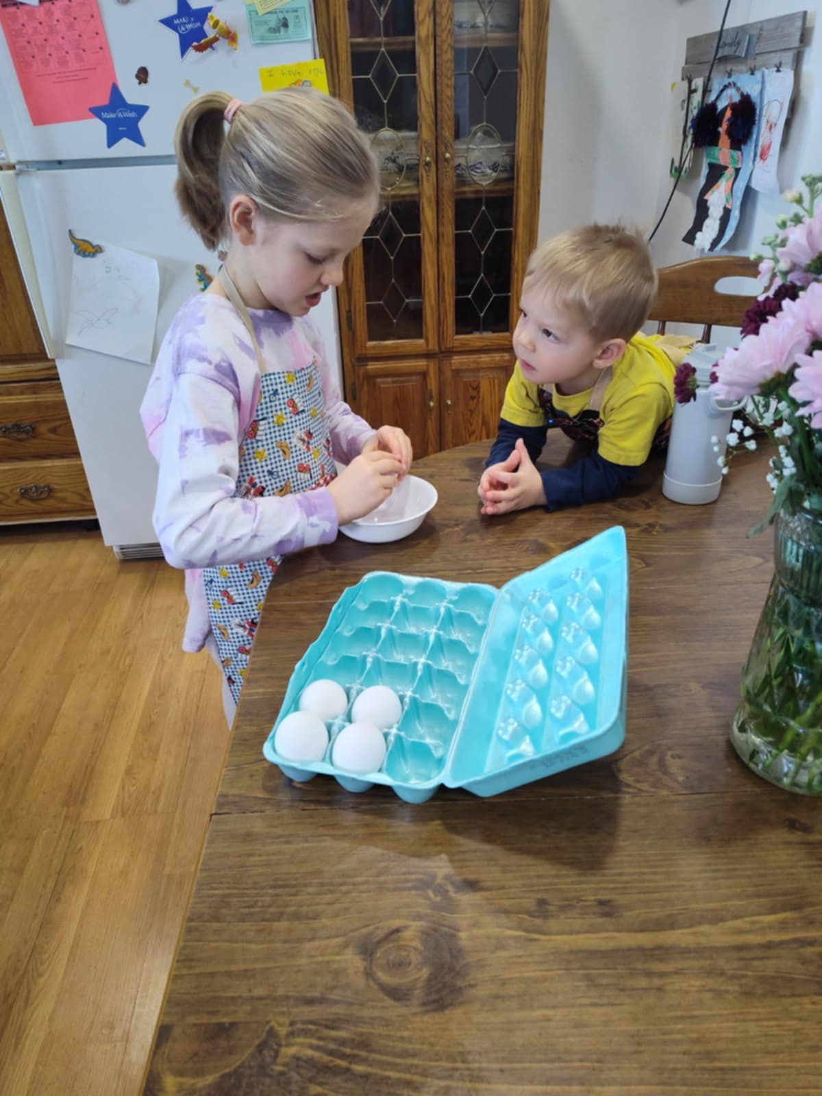 A girl wearing a western style apron  cracking an egg into a bowl with a boy wearing a western style apron  watching.  They are at a wood dinning table with a 18 count egg carton with 4 eggs. The background is a kitchen setting with a fridge and a cabinet.