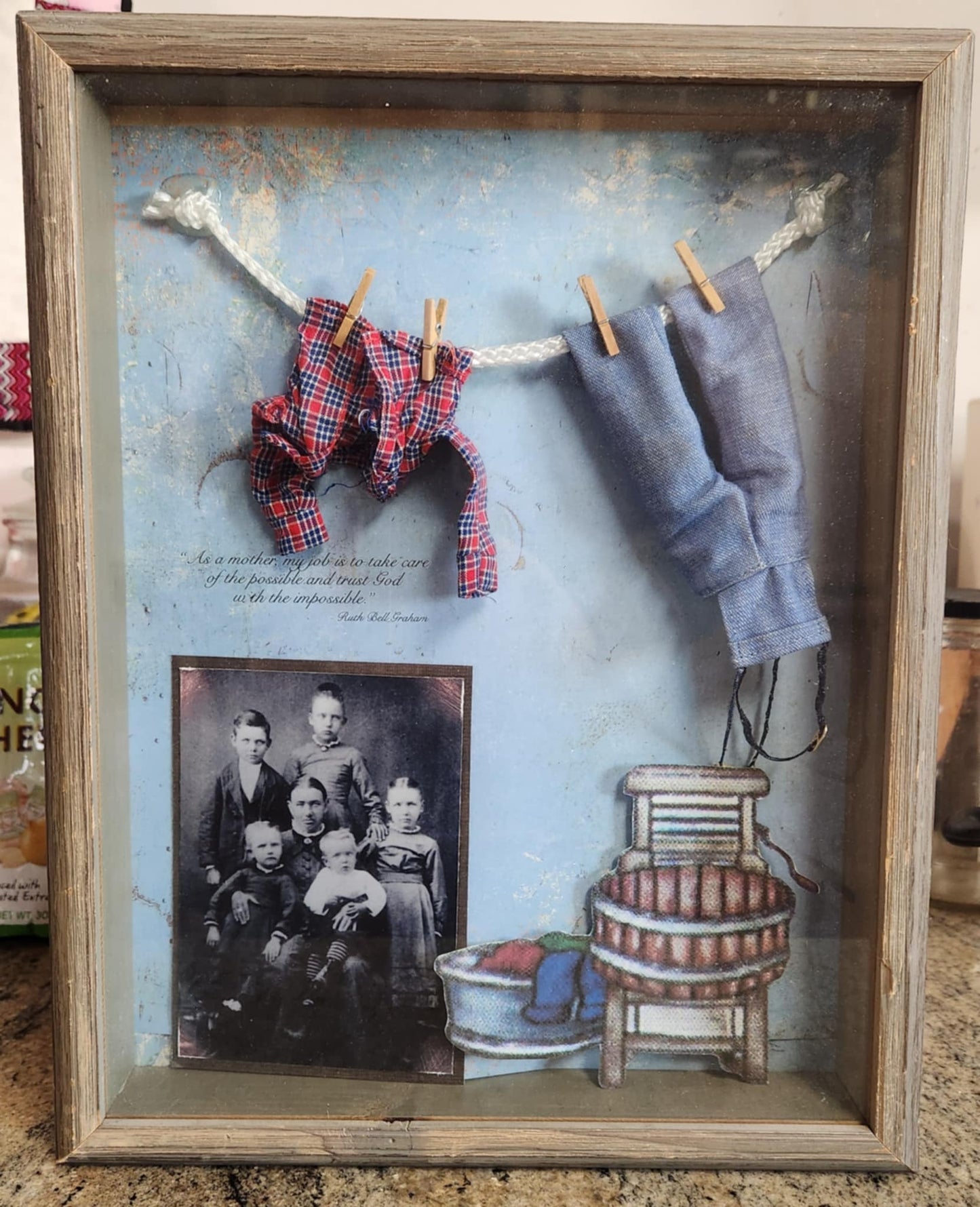 A wood shadow box with a blue background with a laundry line with a shirt and overalls and a photo of a family in the bottom