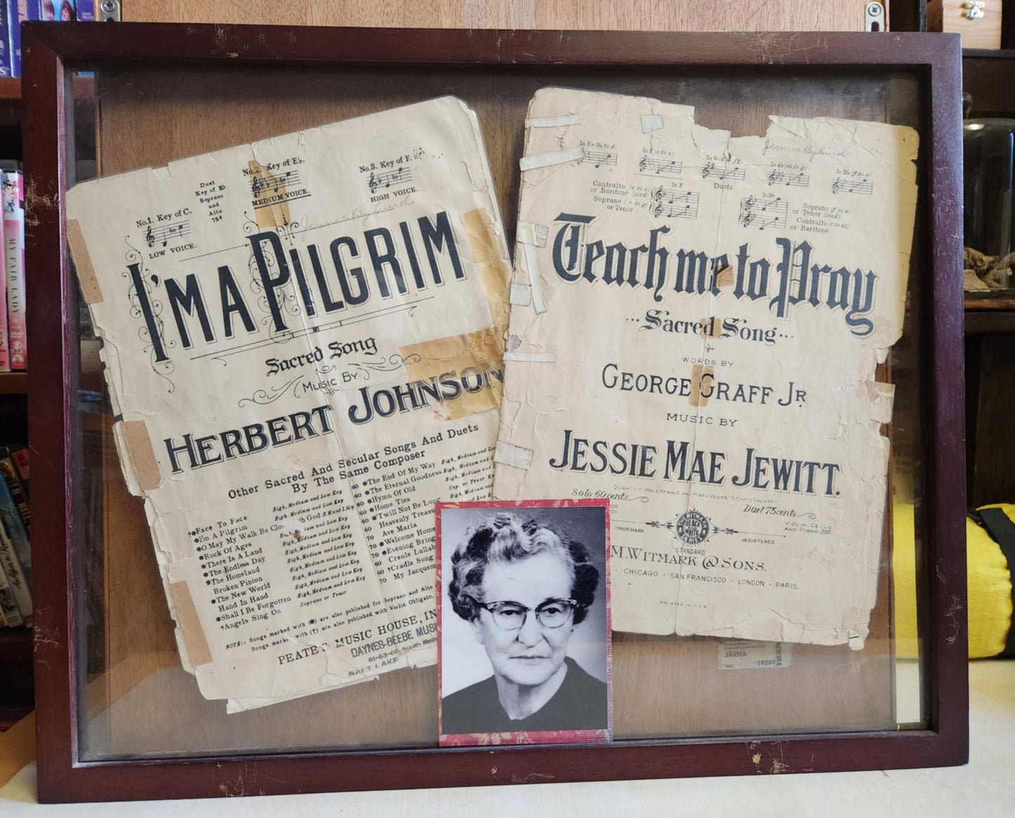 A wood shadow box with two sheet music ones I'm a pilgrim and the other Teach me to pray both look old and warn. There's a photo of a older woman in the center
