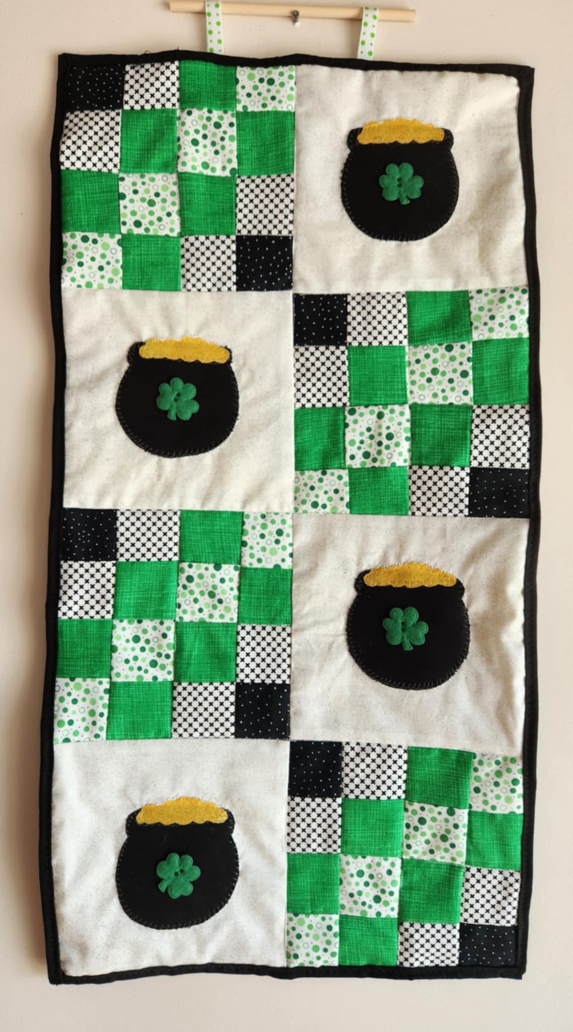 A wall hanging with a pot of gold design, primarily green and black in color, with multiple pots of gold on a white background.