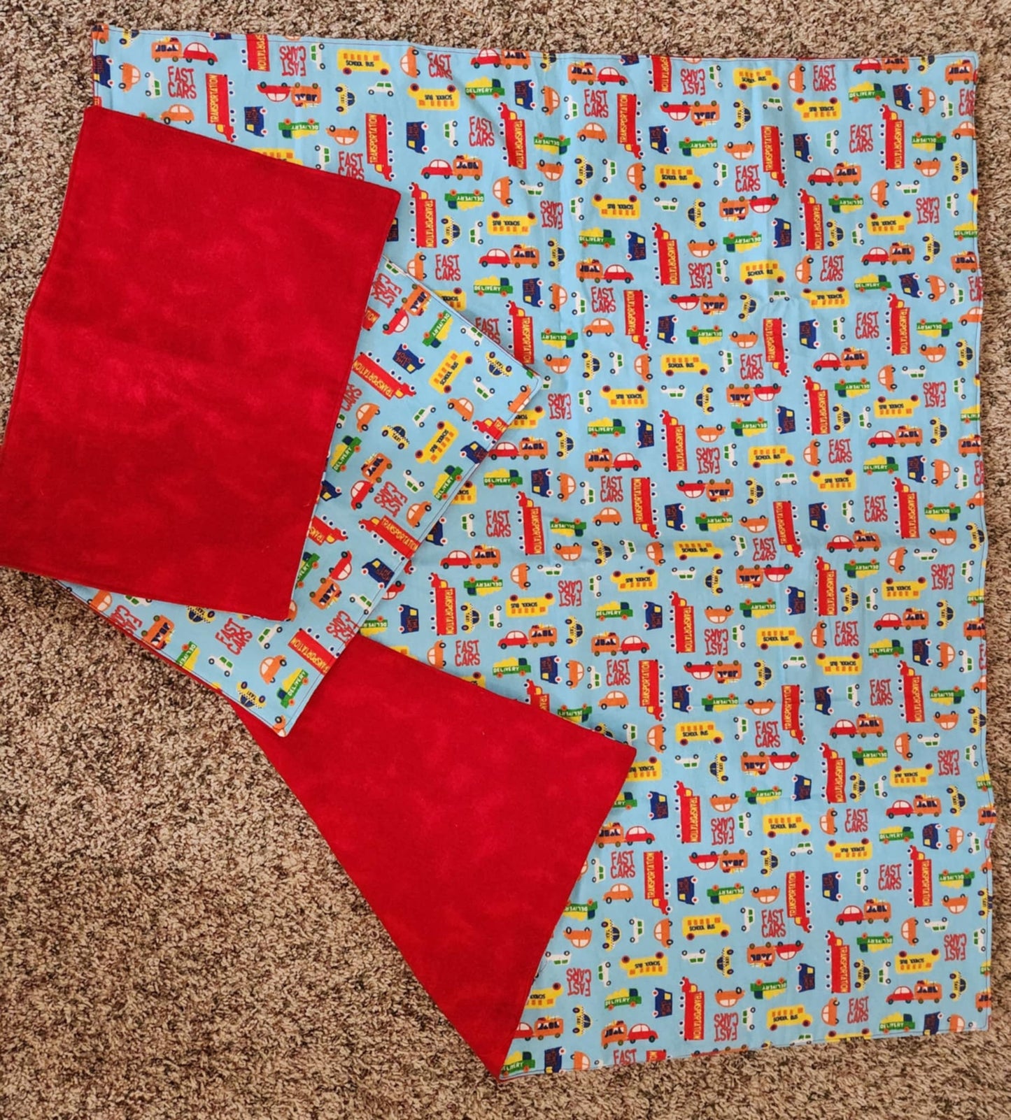 Square Receiving Blanket with Multi Vehicles with a blue background Fleece. With corner turned down showing red background fleece. With two burp rags made from the same fabric on top of the blanket
