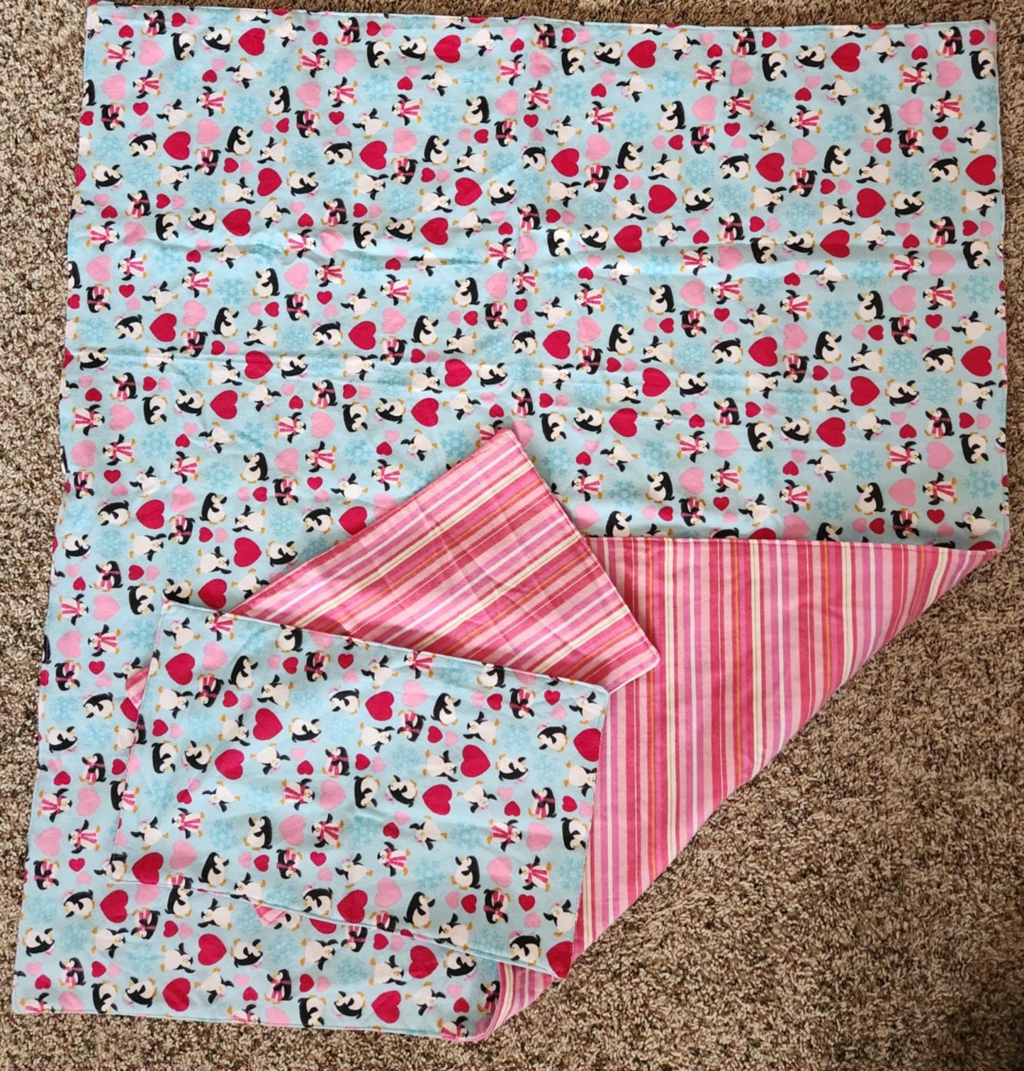 Square Receiving Blanket with Penguins, red and pink hearts with a blue background Fleece. With corner turned down showing pink, red, and white striped background fleece. With two burp rags made from the same fabric on top of the blanket