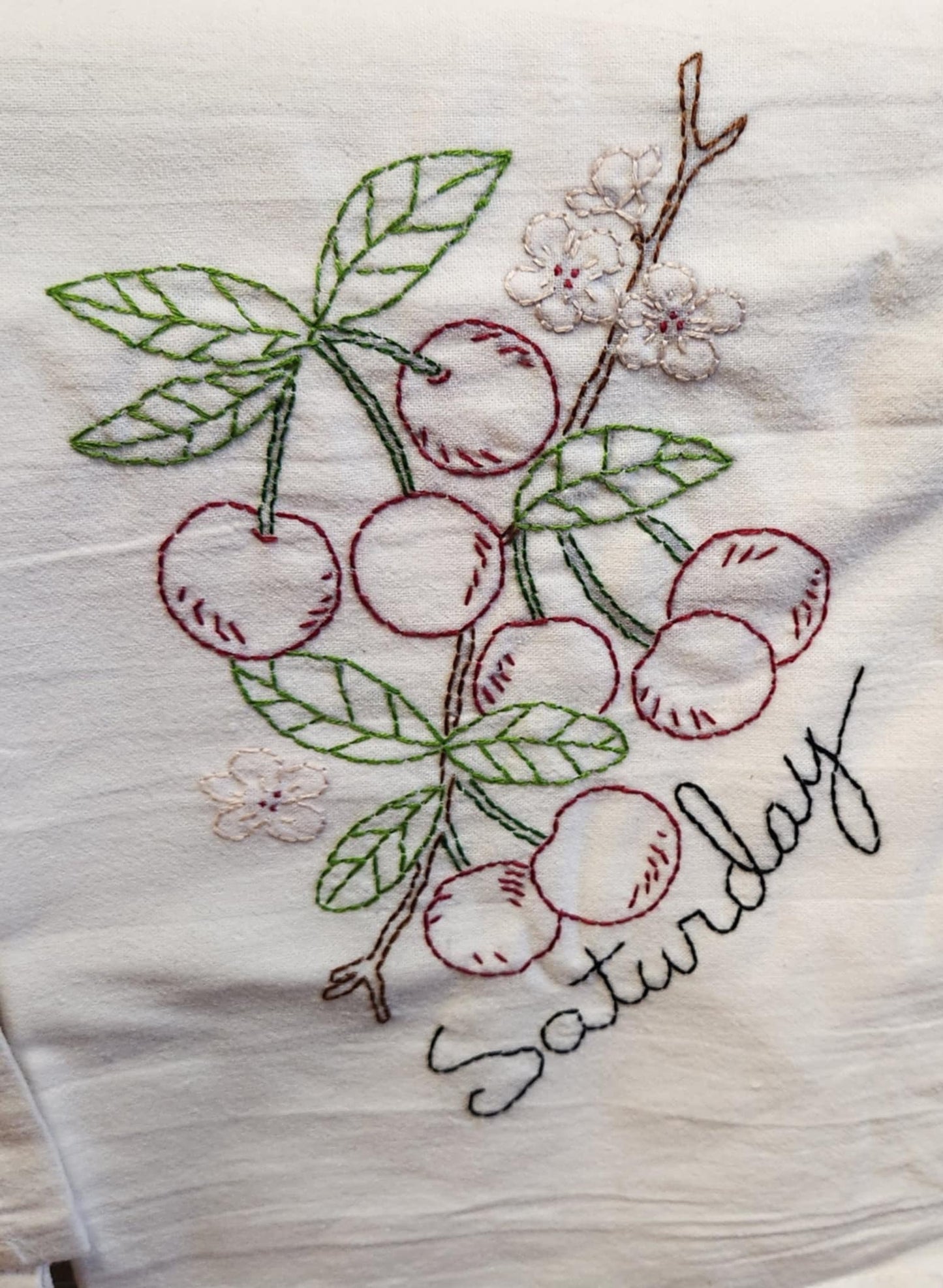 White dish Towel with embroidery with red cherry's with stems attached to a branch with green leaves and small flowers. "Saturday" Embordered in black under the cherry's