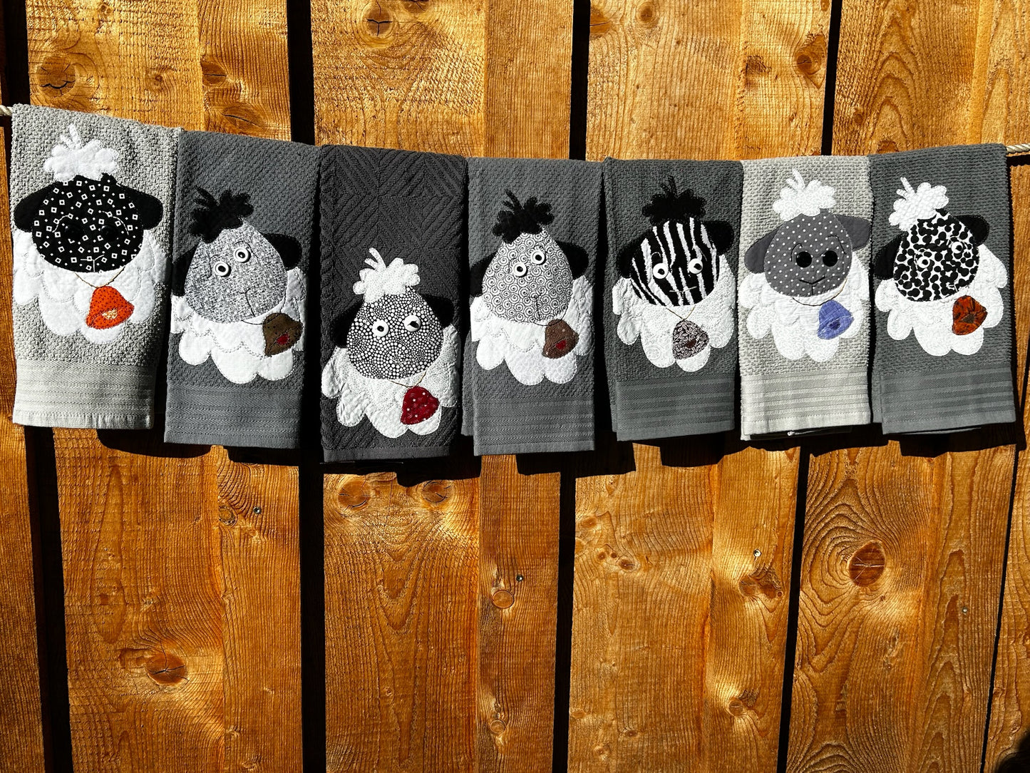 Seven Gray hand towels each a different shade with Sheep on them. Each sheep is ether gray or black. Each Sheep has a bell of different colors and patterns