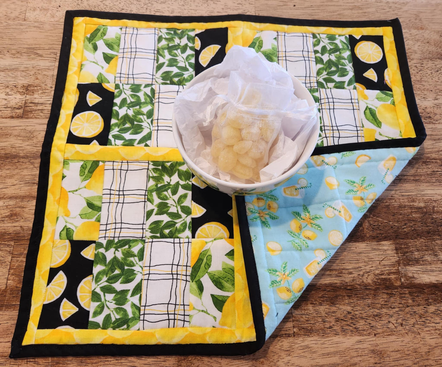 Square Lemon table Runner with black binding, yellow lemons surrounding four square with the pattern of Lemons with green leaves fabric, black background with sliced lemons fabric, green leaves with white background fabric, squiggly black and yellow lines with white back round fabric, repeated twice for each square. The Right corner is folded up showing the backing that has lemons with greens, Glasses with lemonade in them on a blue background.  There is a bowl with lemons and greens on it.
