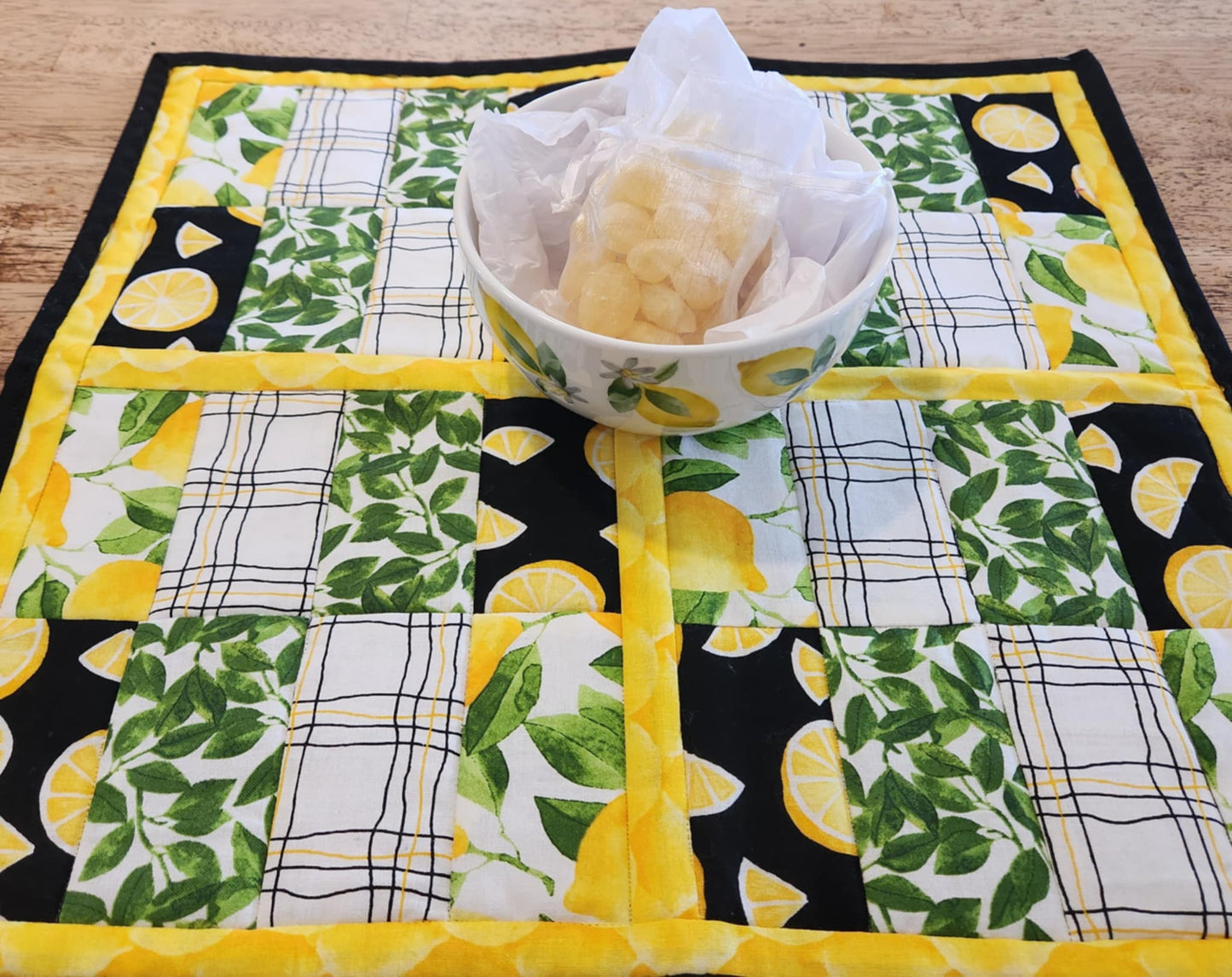 Square Lemon table Runner with black binding, yellow lemons surrounding four square with the pattern of Lemons with green leaves fabric, black background with sliced lemons fabric, green leaves with white background fabric, squiggly black and yellow lines with white back round fabric, repeated twice for each square. There is a bowl with lemons and greens on it.