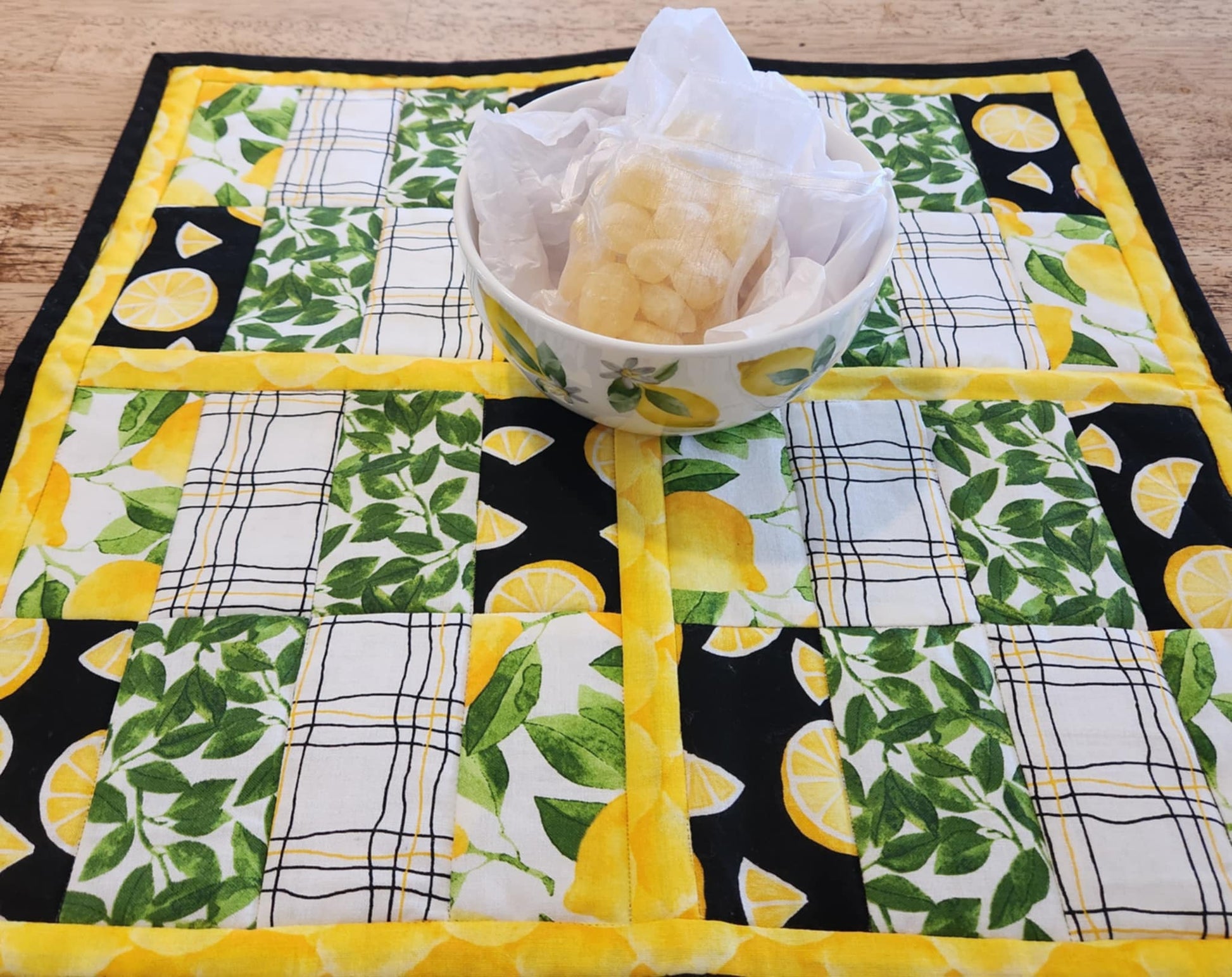 Square Lemon table Runner with black binding, yellow lemons surrounding four square with the pattern of Lemons with green leaves fabric, black background with sliced lemons fabric, green leaves with white background fabric, squiggly black and yellow lines with white back round fabric, repeated twice for each square. There is a bowl with lemons and greens on it.