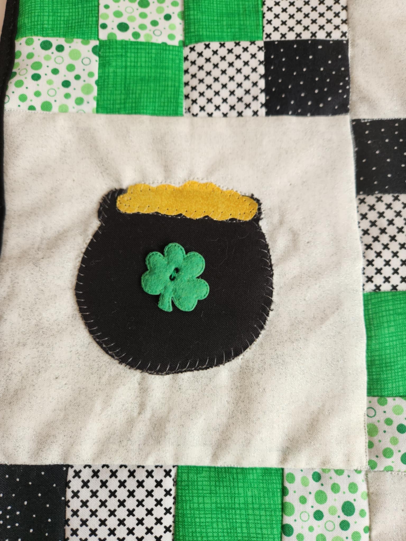 A wall hanging with a pot of gold design for St. Patrick's Day, featuring green and black squares with a yellow pot and a four-leaf clover.