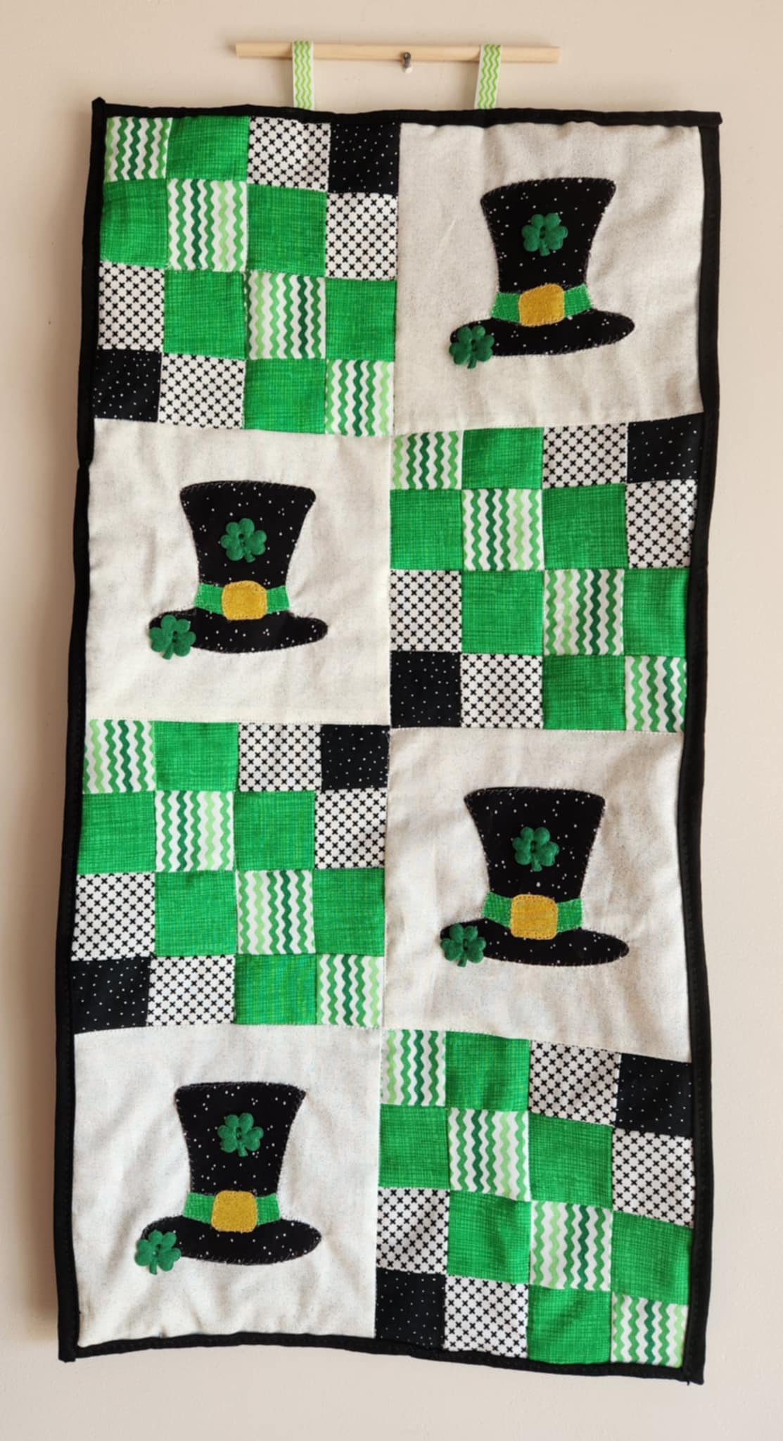 A wall hanging decoration featuring a pattern of green and white squares with embroidered leprechaun hats, with black and gold accents.
