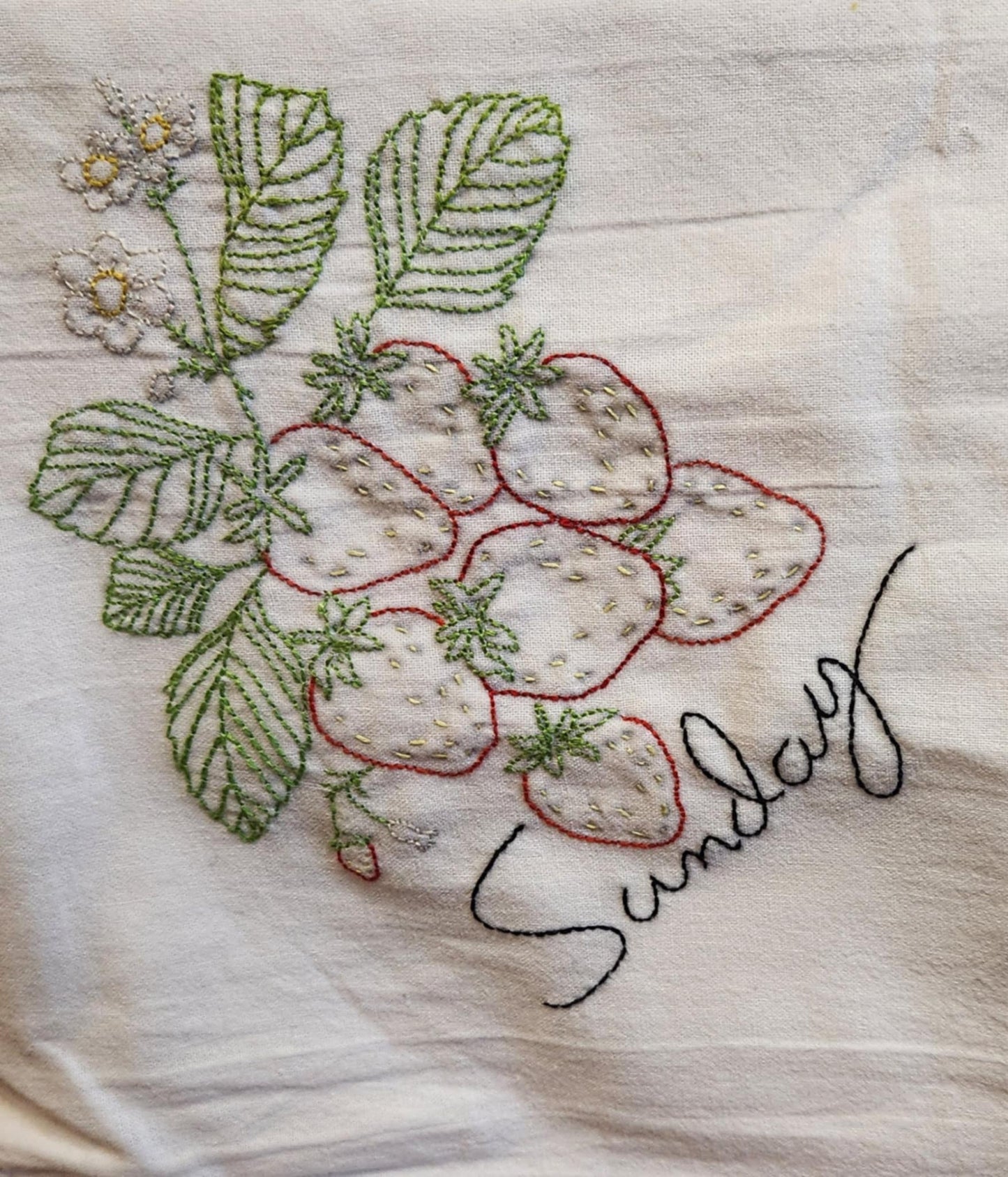 White dish Towel With embordering of a Red Strawberries plant with green leaves and white flowers. "Sunday" embordered under the plant in black thread.