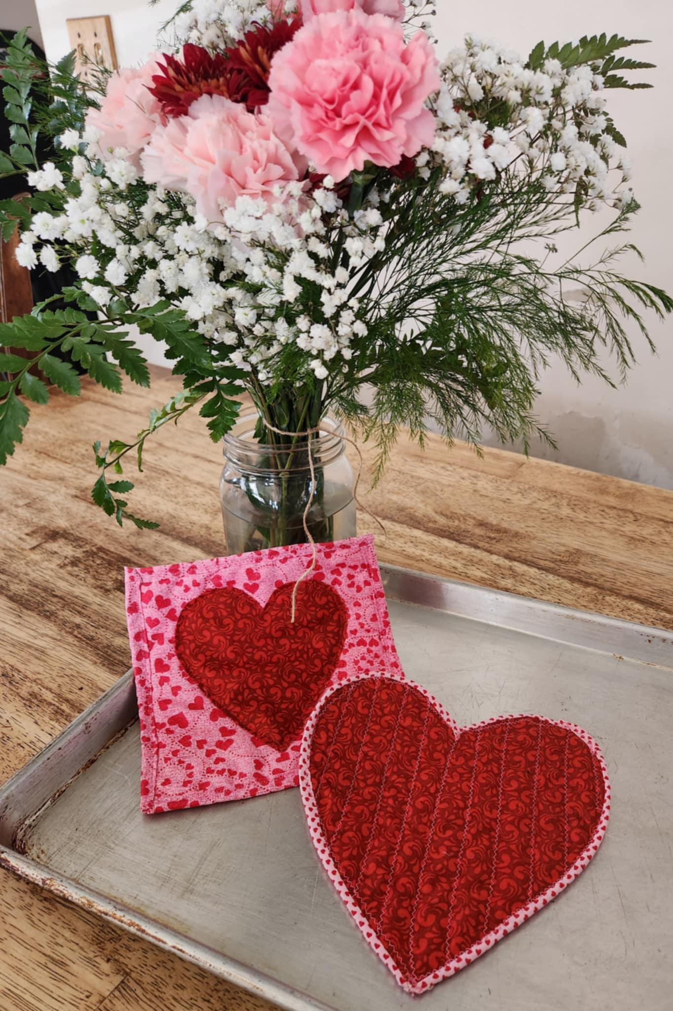 Homemade Valentines Day Heart Hot pads one shaped like a red heart with pink trim and one square hot pad on a pink background and red heart.