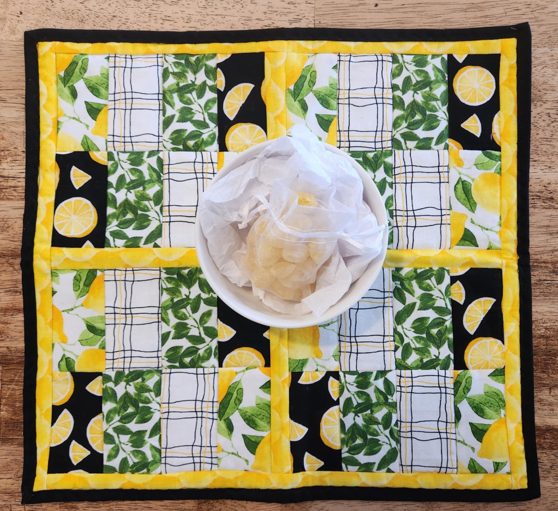Square Lemon table Runner with black binding, yellow lemons surrounding four square with the pattern of Lemons with green leaves fabric, black background with sliced lemons fabric, green leaves with white background fabric, squiggly black and yellow lines with white back round fabric, repeated twice for each square. There is a bowl with lemons and greens on it.
