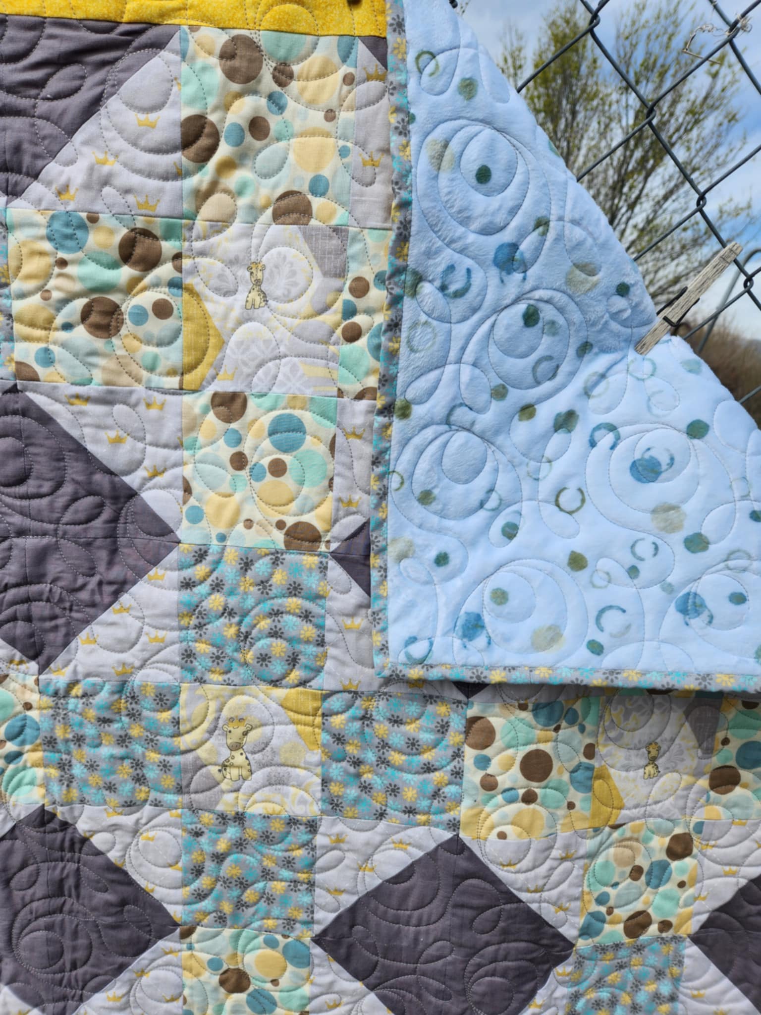 A Quilt With a octagon patters with squares of circles in brown, blue and yellow, rectangles of solid dark gray and light gray background with gold crowns and a square with a giraffe in the center surrounded by gray and gold octagons. The top right corner is turned down showing the Light blue background with full circles and half circles on minky fabric.