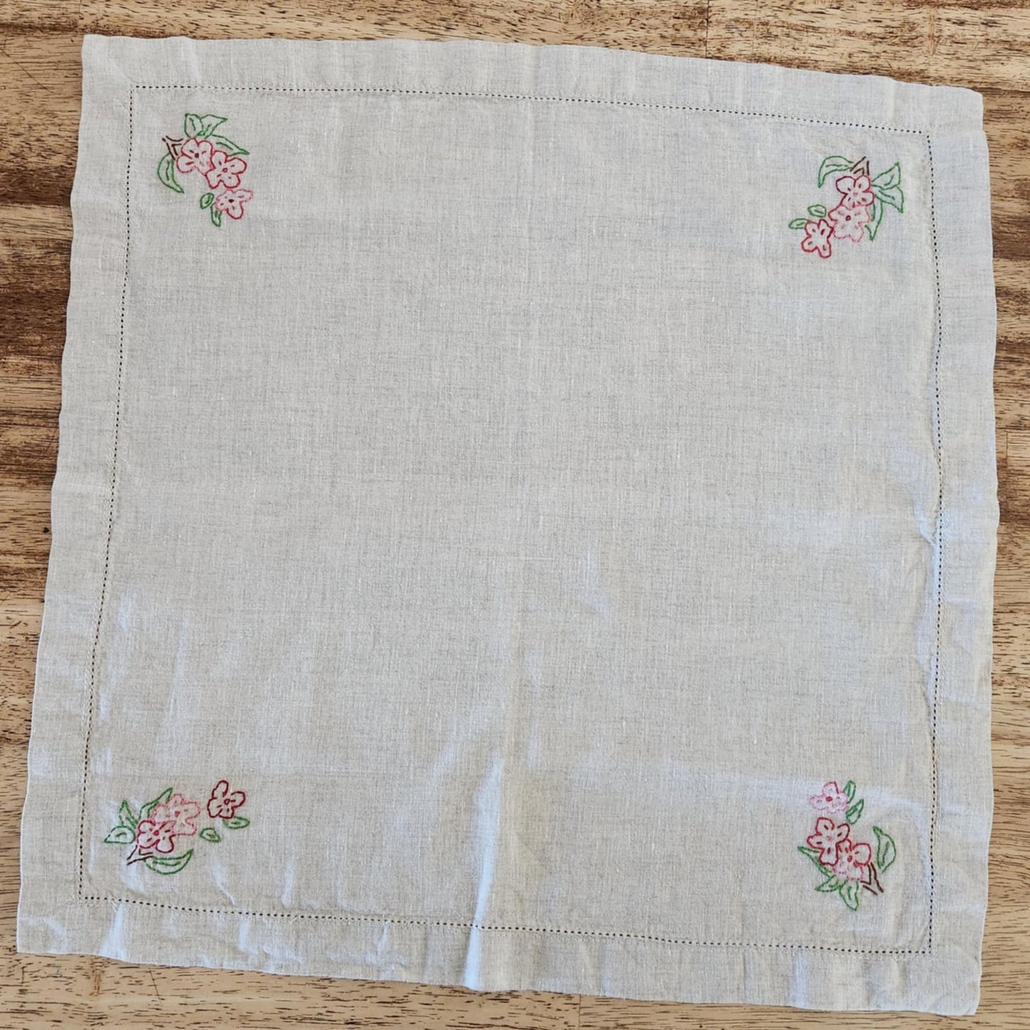 White cotton square table topper with a border of small dots and pink, purple and yellow flowers and green leaf's in each corner