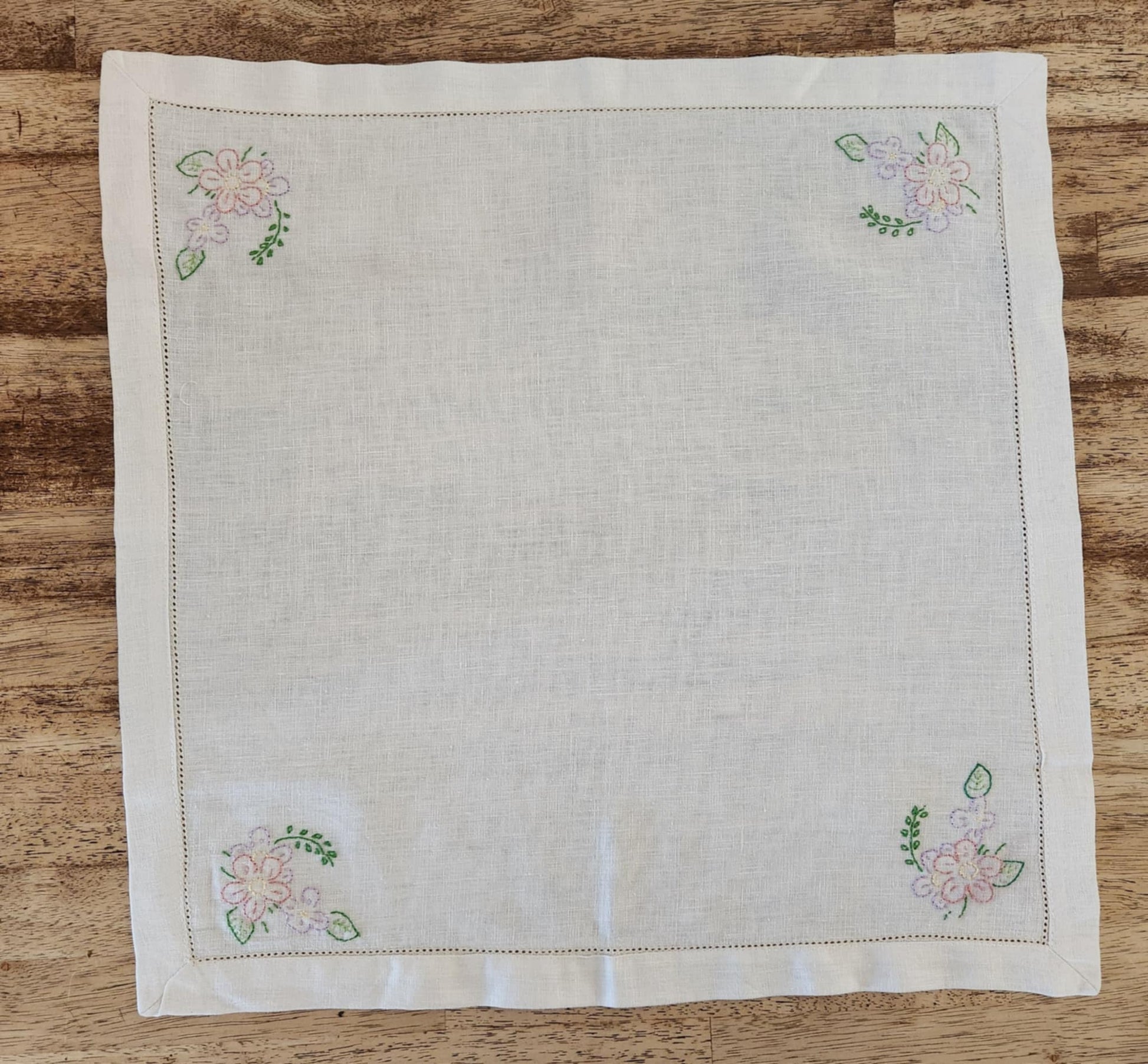White cotton square table topper with a border of small dots and pink, purple and yellow flowers and green leaf's in each corner