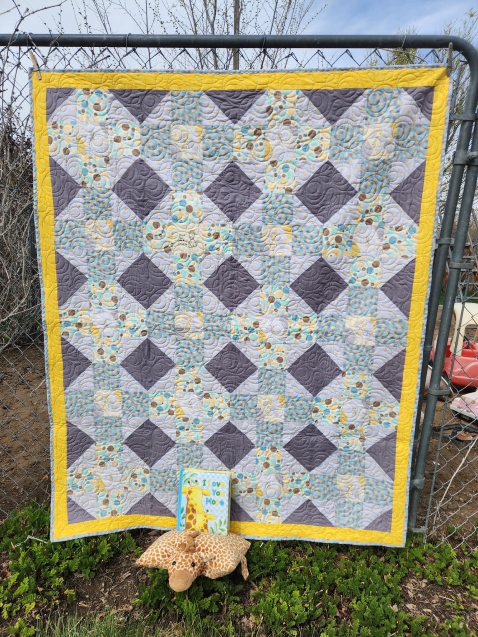 A Quilt With a octagon patters with squares of circles in brown, blue and yellow, rectangles of solid dark gray and light gray background with gold crowns and a square with a giraffe in the center surrounded by gray and gold octagons. at the bottom is a stuff giraffe and a book called i love you more