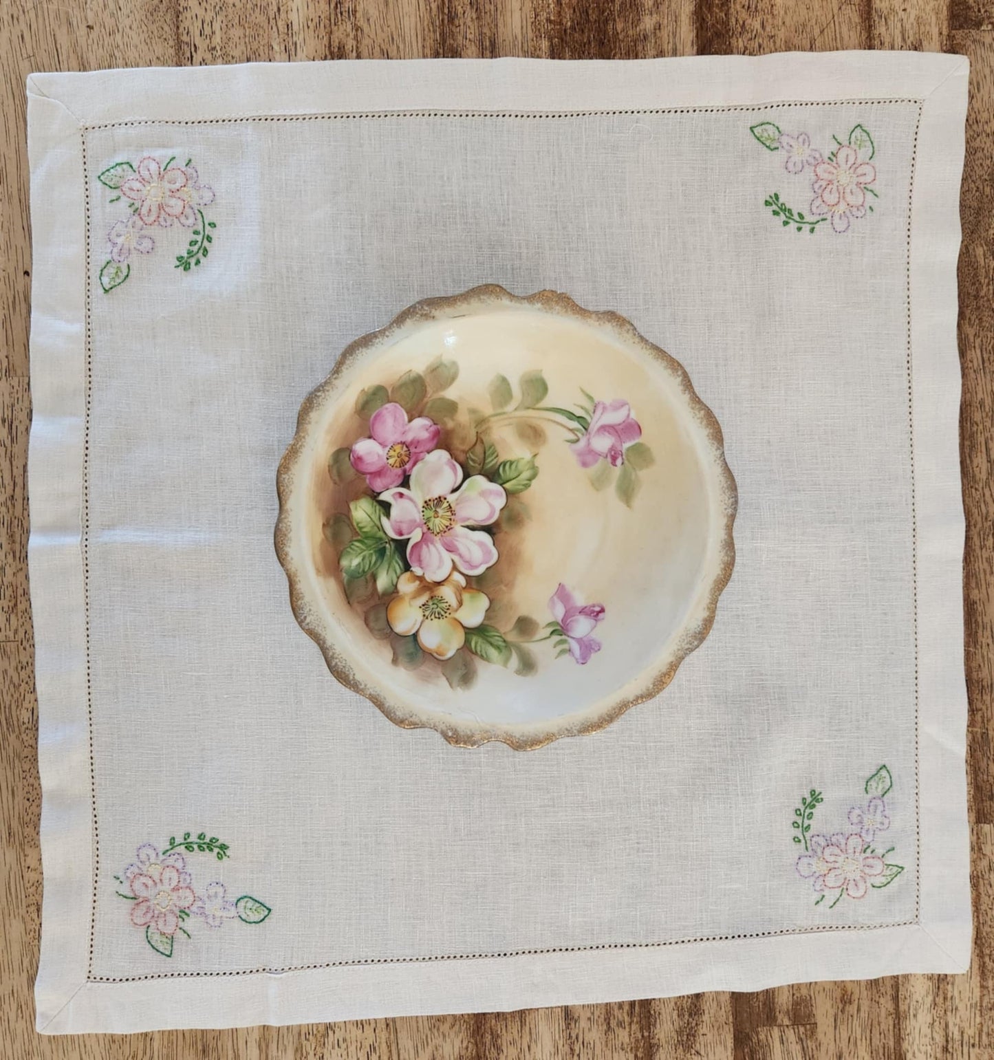 White cotton square table topper with a border of small dots and pink, purple and yellow flowers and green leaf's in each corner
