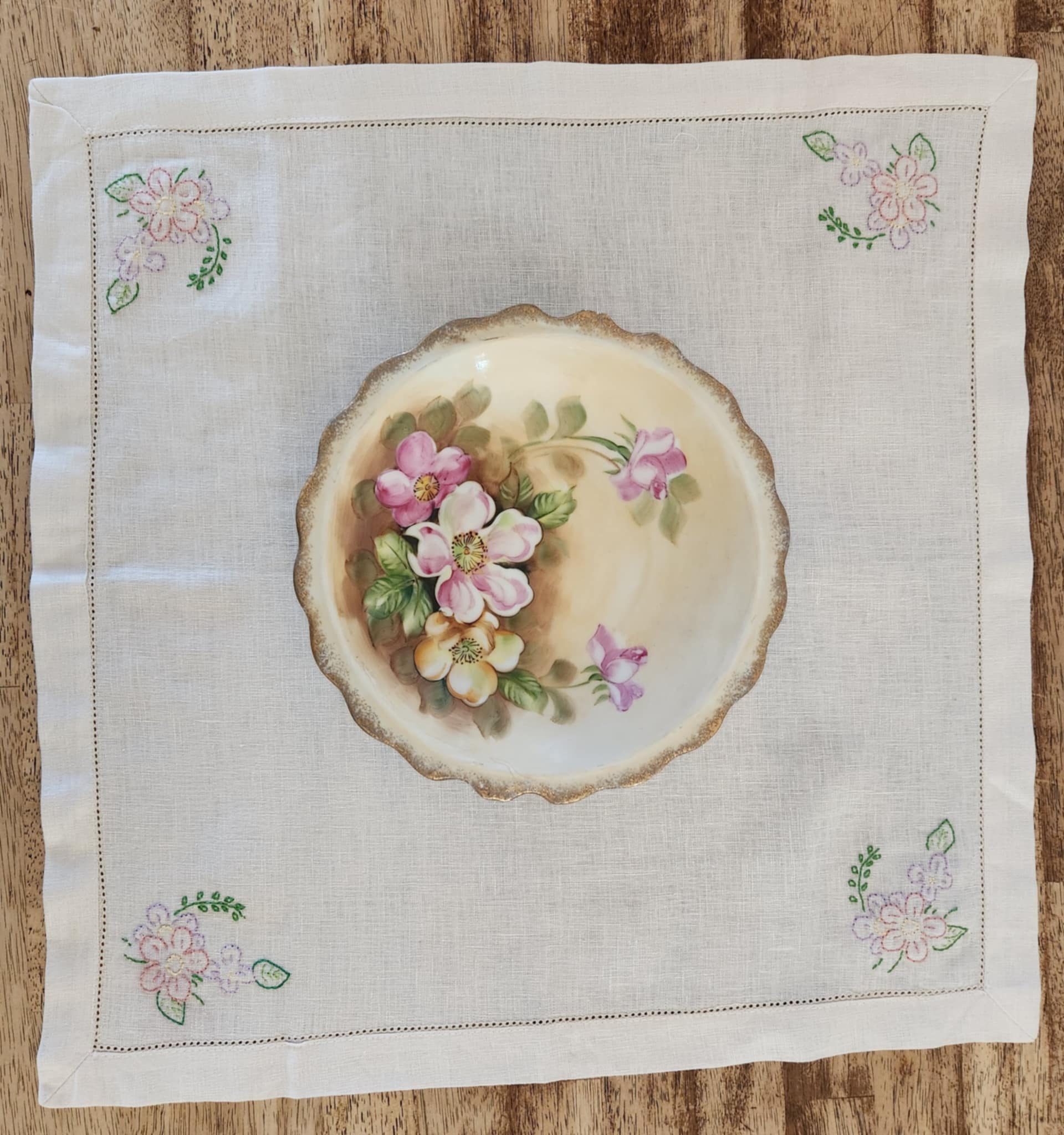 White cotton square table topper with a border of small dots and pink, purple and yellow flowers and green leaf's in each corner