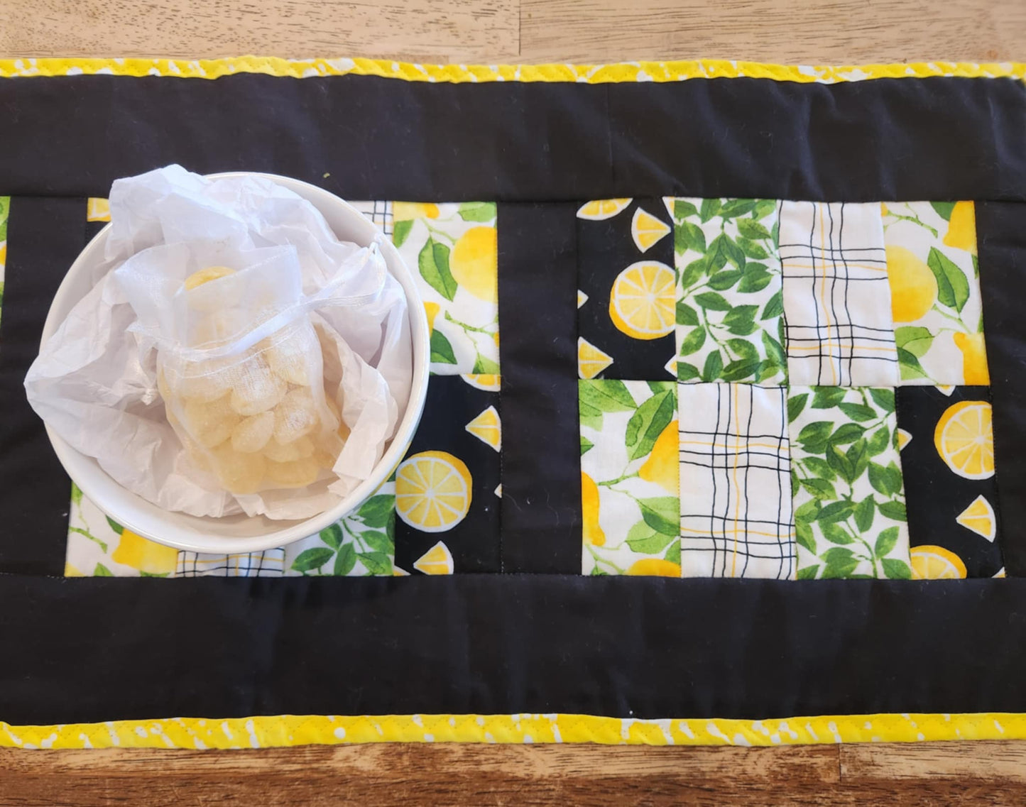 Close up of table runner with Yellow and white binding, with black surrounding four square with the pattern of Lemons with green leaves fabric, black background with sliced lemons fabric, green leaves with white background fabric, squiggly black and yellow lines with white back round fabric, repeated twice for each square. There is a bowl with lemons and greens on it.