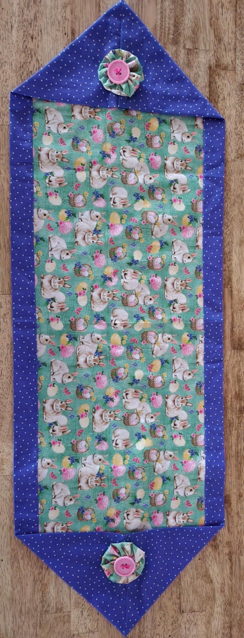A top view table runner with bunnies' with baskets filled with Easter eggs  in the middle surrounded with a purple poke-a-dots outside with a pink button on eachside