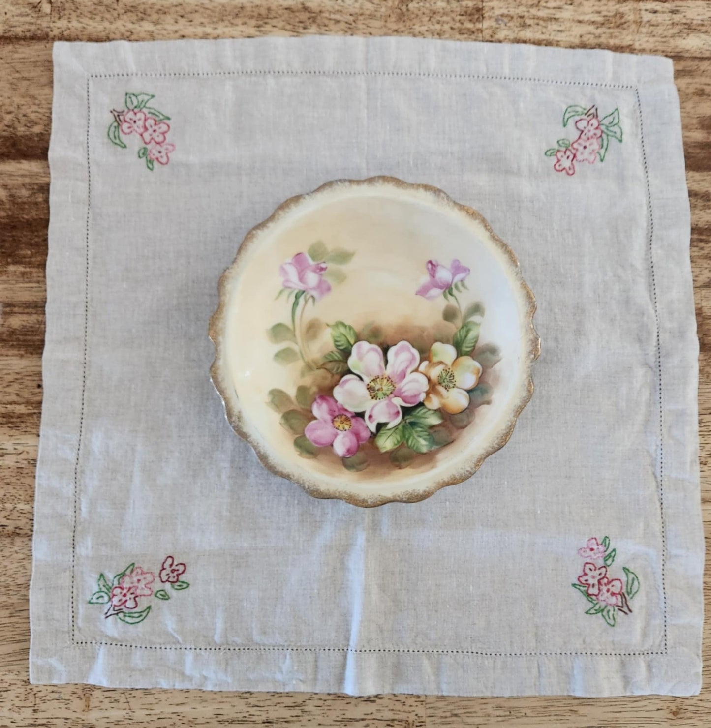 White cotton square table topper with a border of small dots and pink, purple and yellow flowers and green leaf's in each corner