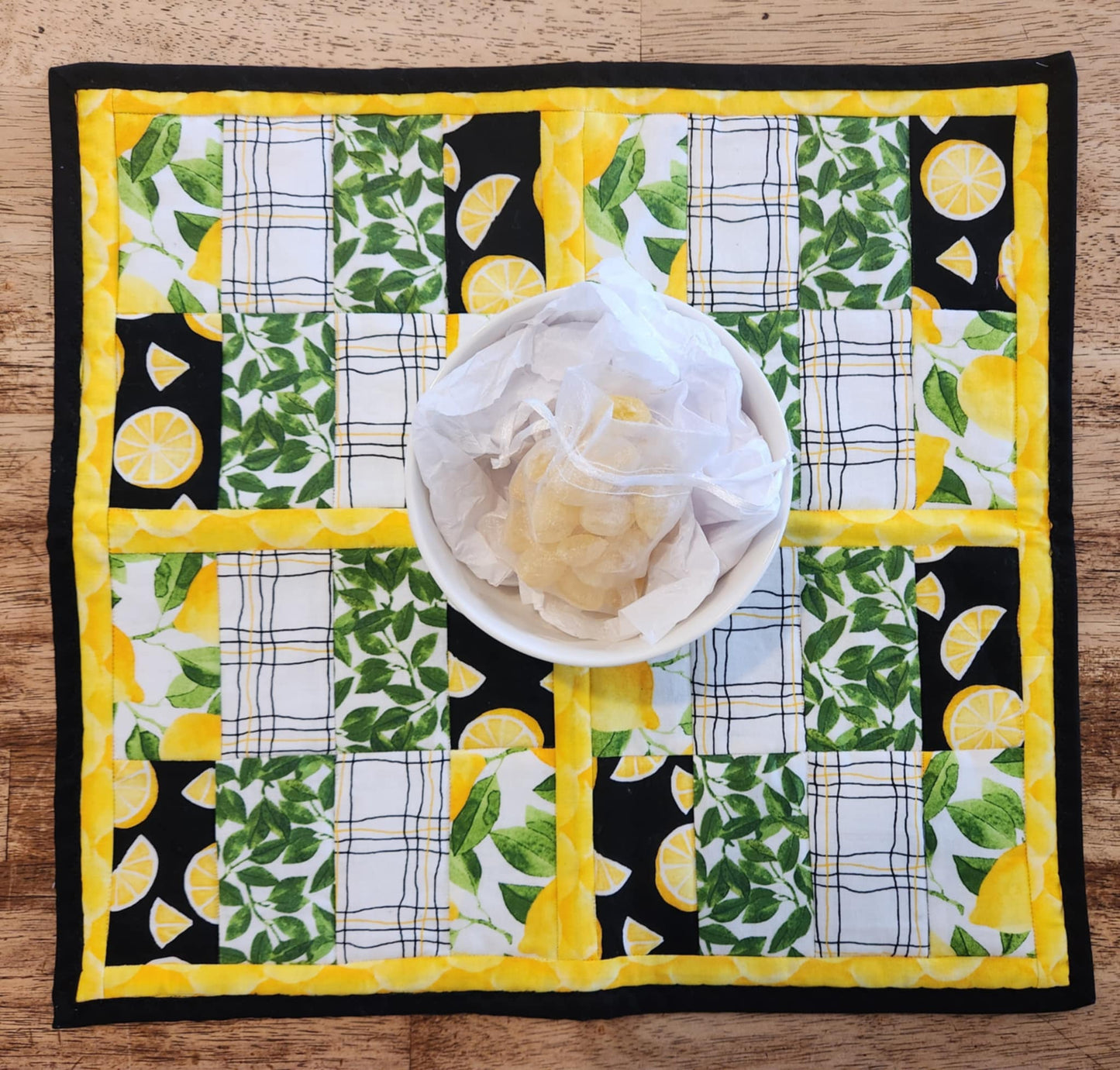 Square Lemon table Runner with black binding, yellow lemons surrounding four square with the pattern of Lemons with green leaves fabric, black background with sliced lemons fabric, green leaves with white background fabric, squiggly black and yellow lines with white back round fabric, repeated twice for each square. There is a bowl with lemons and greens on it.