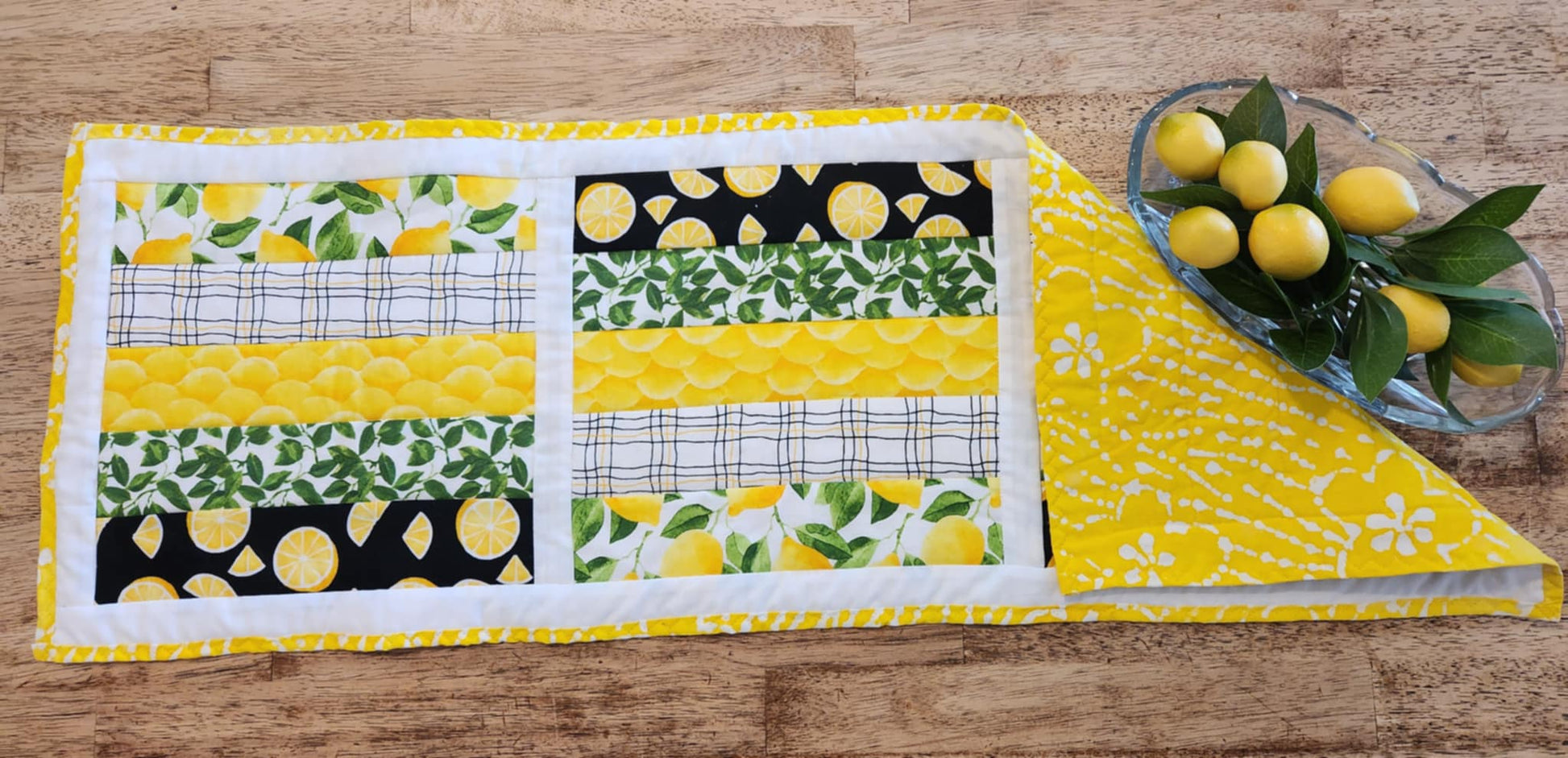 A table runner with Yellow and white binding, with white surrounding three square with the pattern of Lemons with green leaves fabric, black background with sliced lemons fabric, green leaves with white background fabric, squiggly black and yellow lines with white back round fabric, in strips for each square. The right corner is turned down showing yellow fabric with white flowers. There is a glass oval bowl with fake lemons and greens in it.
