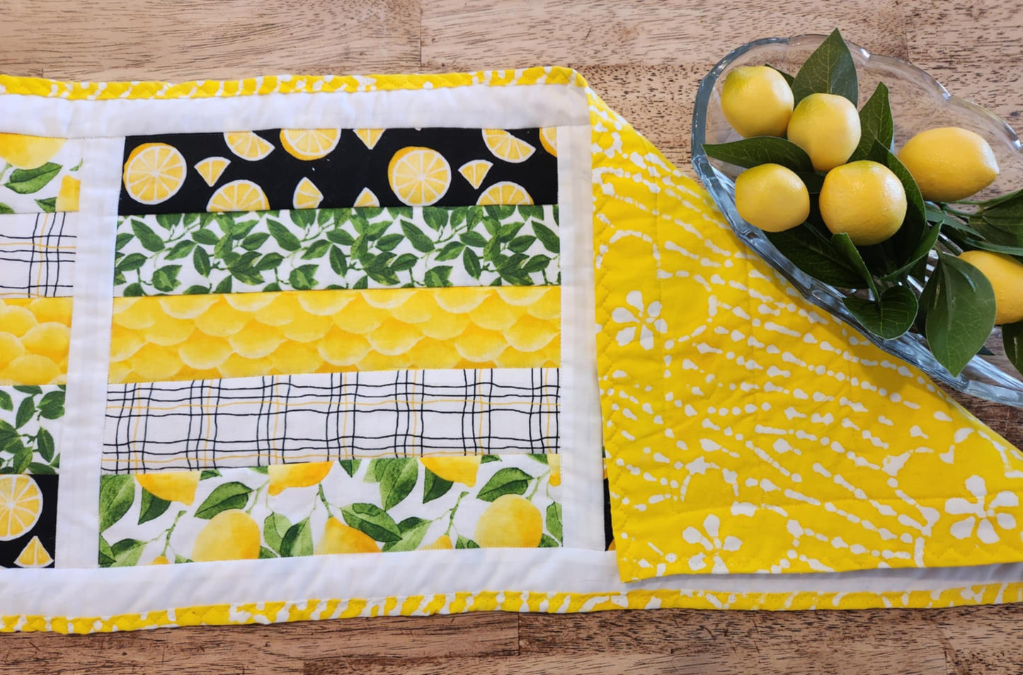 Close up of table runner with Yellow and white binding, with white surrounding three square with the pattern of Lemons with green leaves fabric, black background with sliced lemons fabric, green leaves with white background fabric, squiggly black and yellow lines with white back round fabric, in Strips for each square. The right corner is turned down showing yellow fabric with white flowers. There is a glass oval bowl with fake lemons and greens in it.