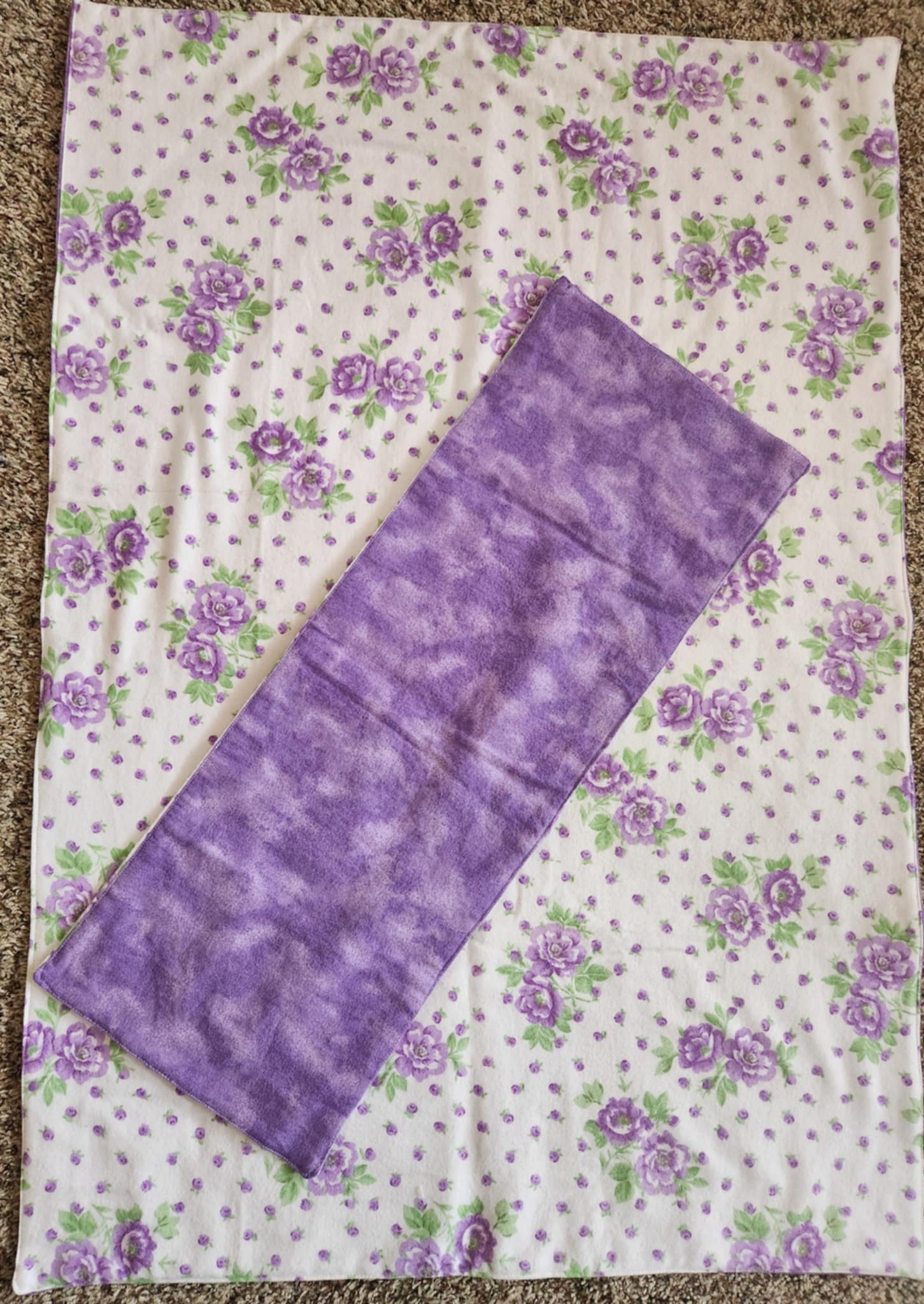 A purple and green floral-patterned receiving blanket with a solid purple burp rag laid on top of it. 