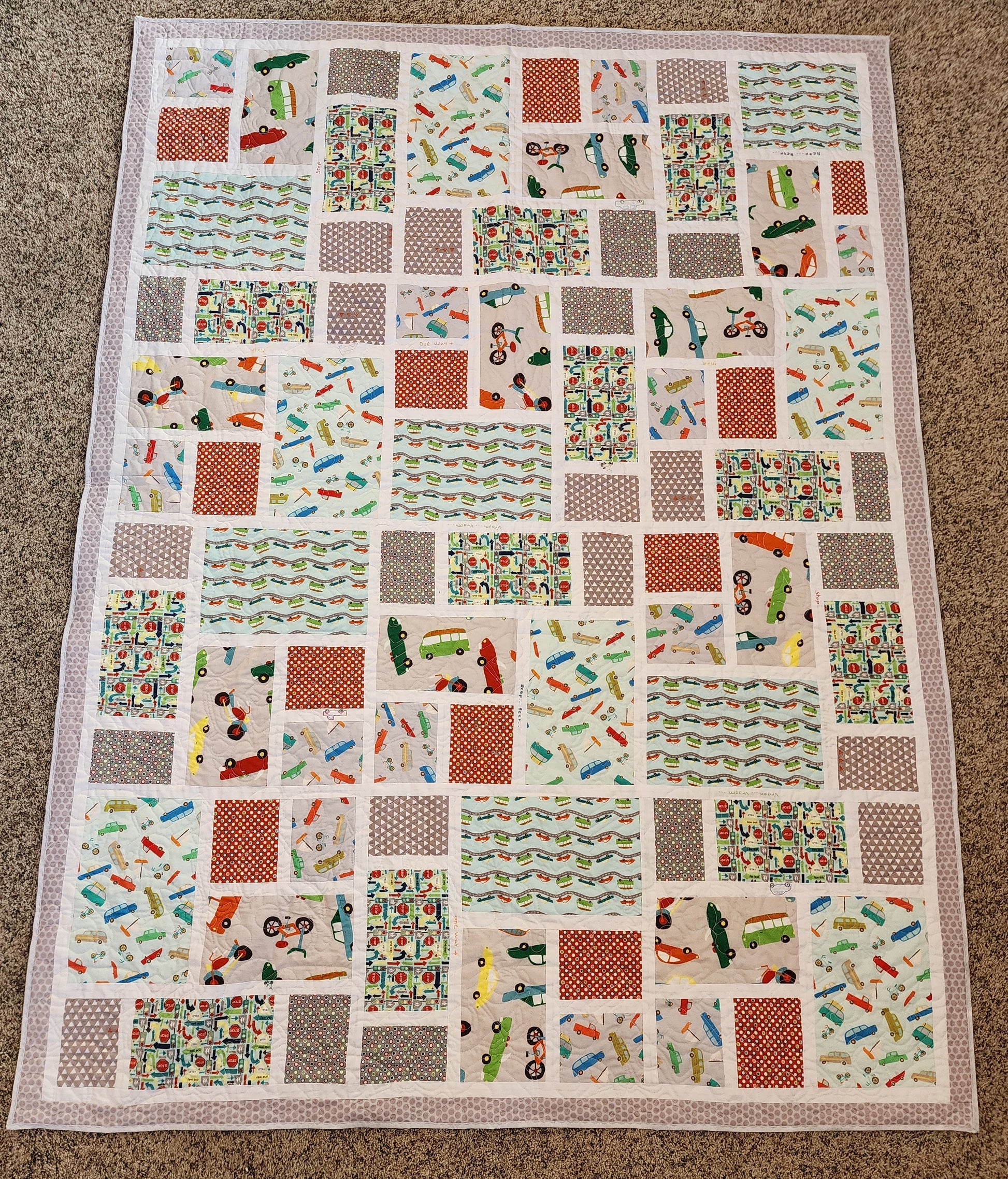 The image shows a patchwork storybook quilt. The quilt is made up of numerous squares of fabric, each with a different design featuring various vehicles such as cars, trucks, and bicycles. The overall color scheme is muted, with colors like light grey, light teal, red, and burnt orange.  The squares are arranged in a seemingly random but aesthetically pleasing pattern. The quilt appears to be handmade and displays good craftsmanship.