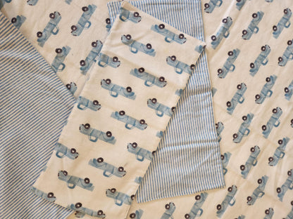 Close up on Receiving Blanket with Baby blue Pick up trucks with white background Fleece. With corner turned down showing baby blue and white Strip fleece. With tow burp rags  made from the same fabric on top of the blanket