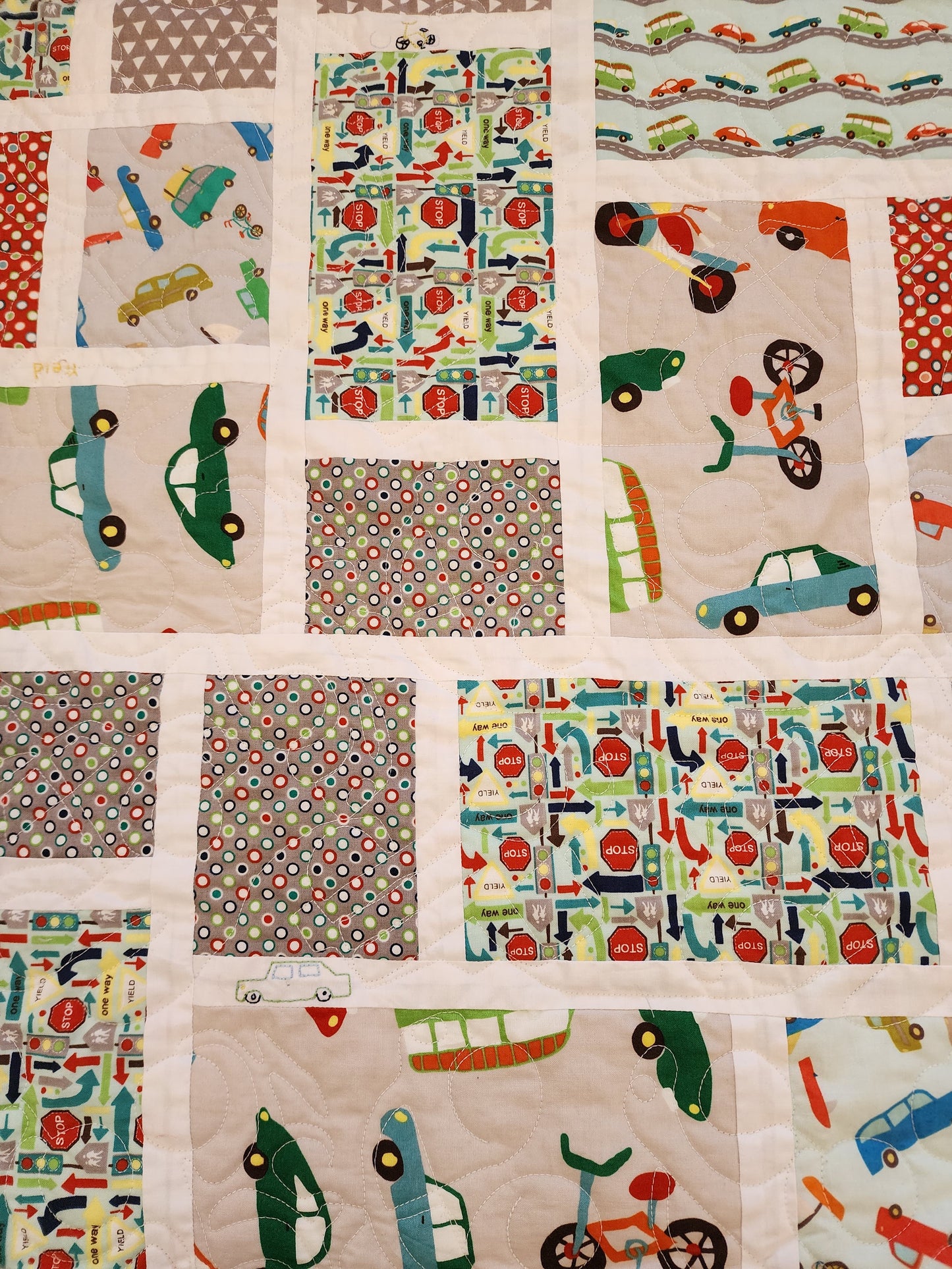 The image shows a close up on a  patchwork storybook quilt. The car quilt is made up of numerous squares of fabric, each with a different design featuring various vehicles such as cars, trucks, and bicycles. The overall color scheme are muted, with colors like light grey, light teal, and burnt orange.  The squares are arranged in a seemingly random but aesthetically pleasing pattern. The car quilt appears to be handmade and displays good craftsmanship.