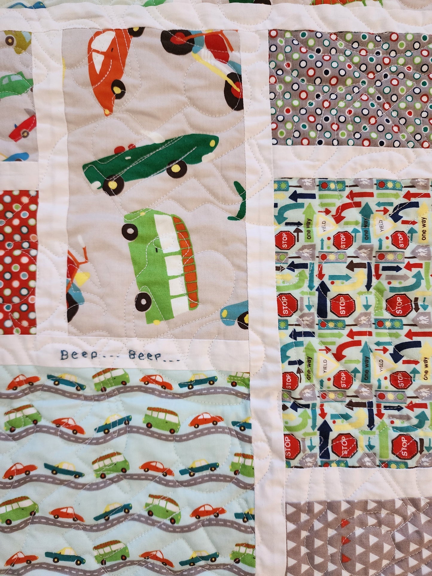 The image shows a detail of a patchwork quilt made of various fabrics. The quilt features a playful transportation theme, with different patterns depicting cars, buses, traffic signs, arrows indicating directions, and simple geometric patterns like polka dots and triangles.  The fabrics are sewn together in a grid-like pattern, and some sections have additional quilting stitches creating a wavy, road-like design.  The words "Beep... Beep..." are embroidered onto one of the sections.