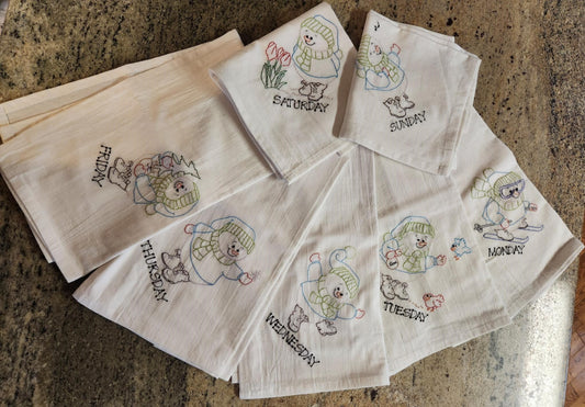 A set of beige flour sack dish towels with embroidered snowman designs and text indicating days of the week. spread out to see each of the on a granite background 